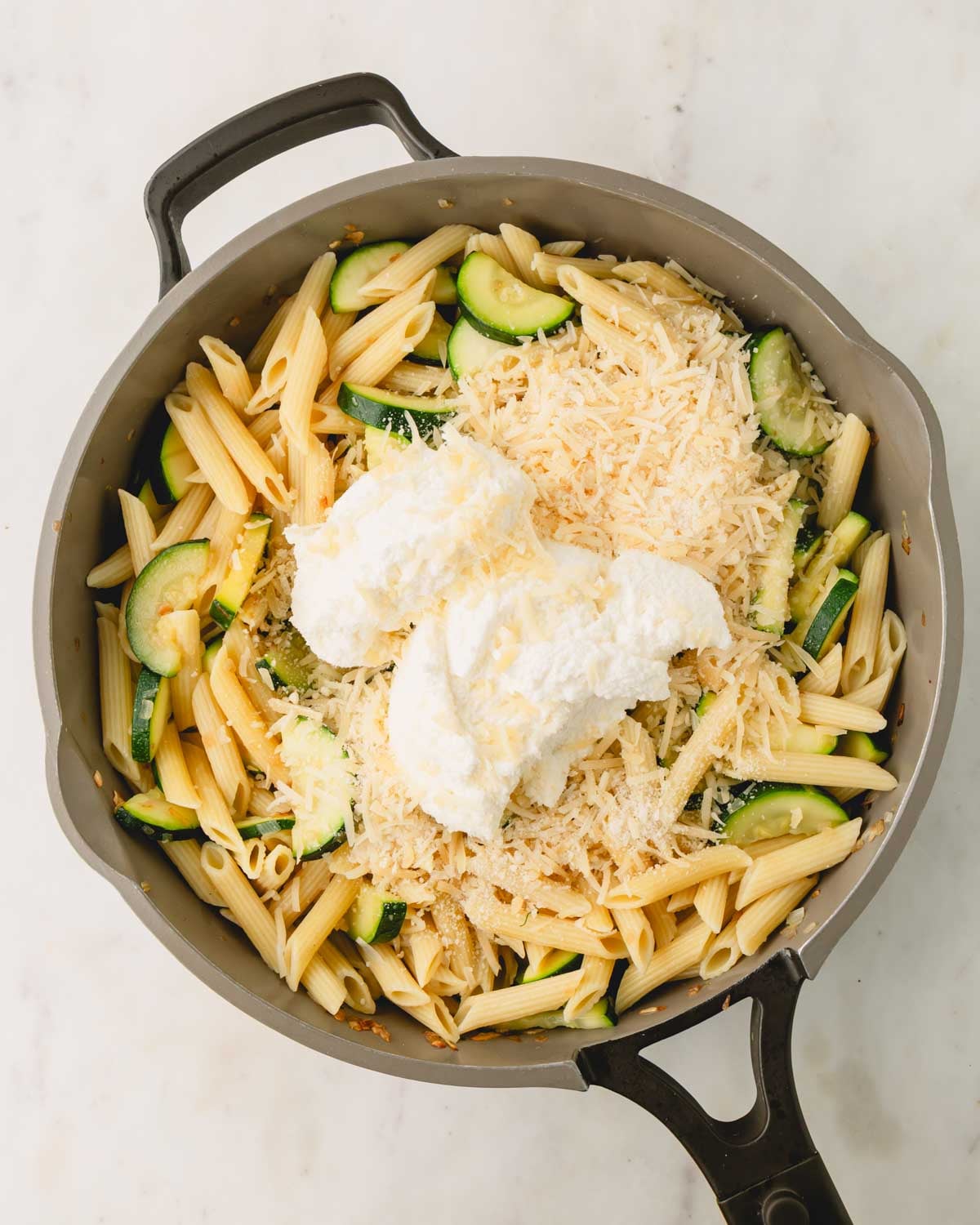Ricotta and parmesan cheese are added to a large skillet with penne pasta and zucchini.