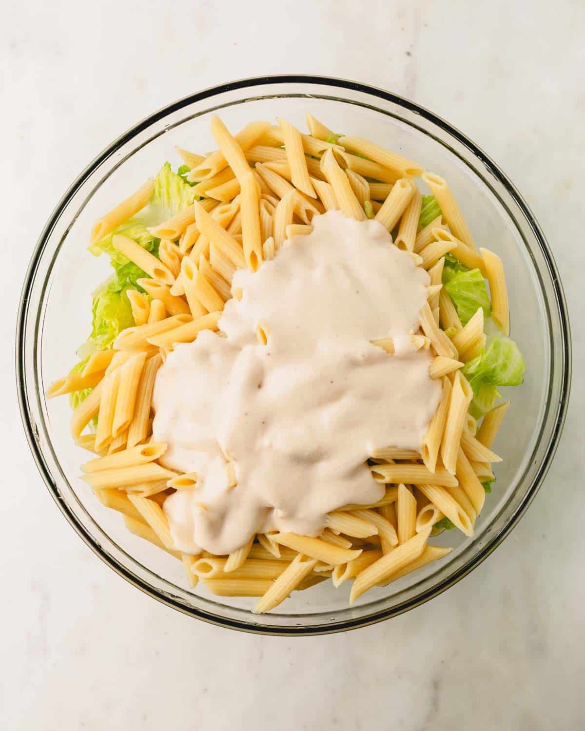 The Caesar pasta ingredients in a large glass bowl with Caesar dressing on top.