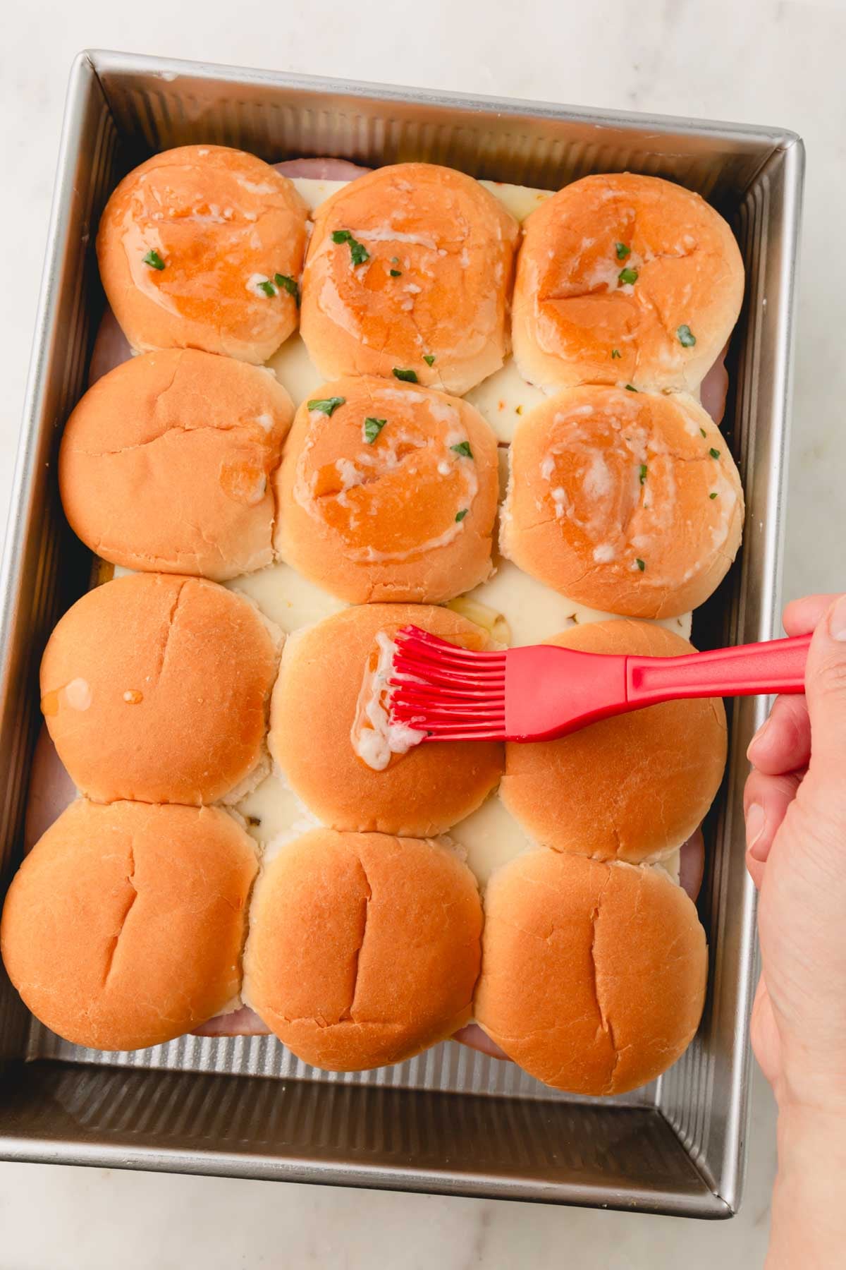A hand is holding a red brush that is brushing on the melted garlic butter on top o the slider rolls.