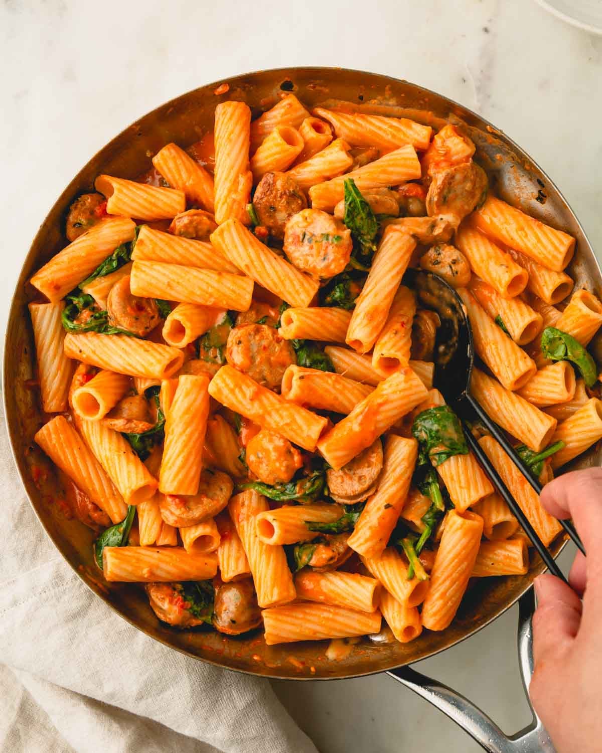 Chicken sausage and pasta recipe in a skillet with a hand scooping some with a large spoon.