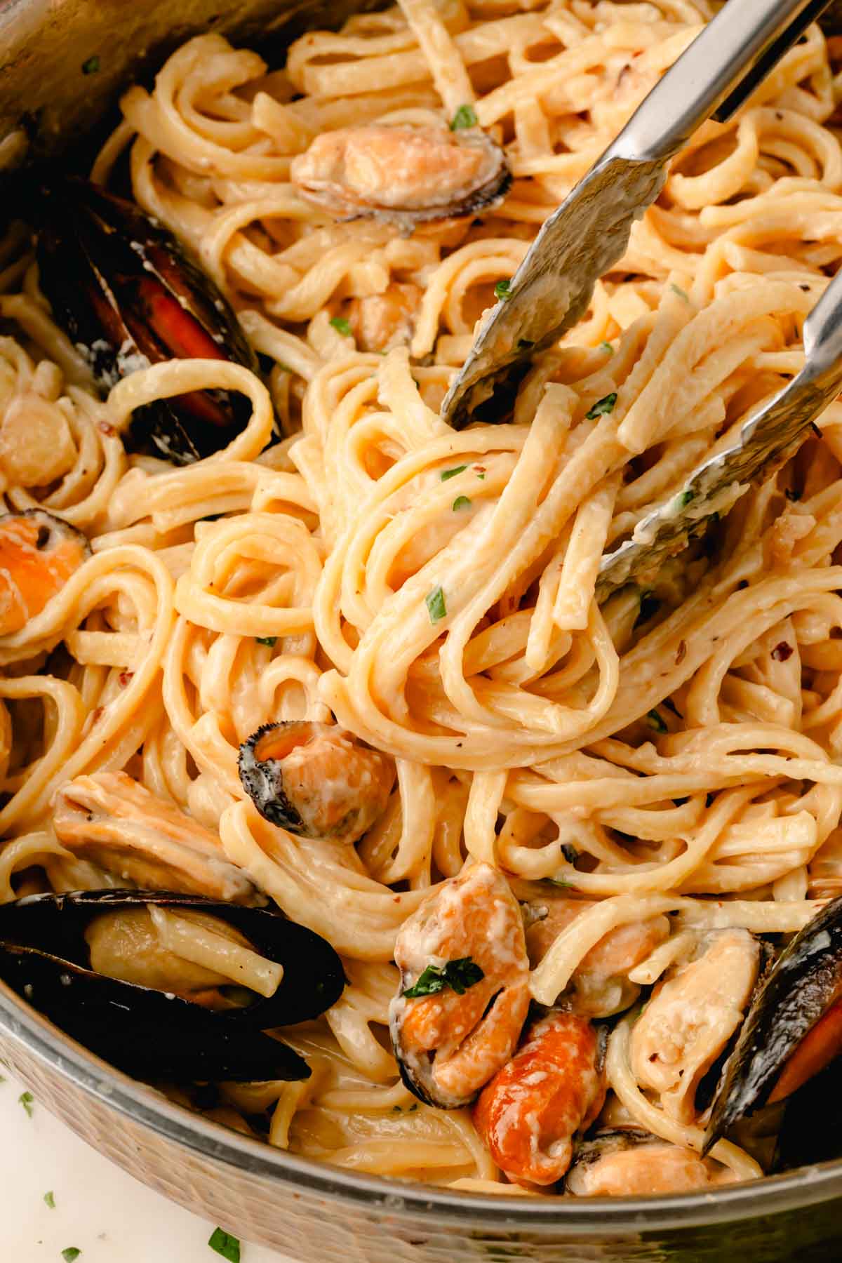 Tongs are tossing the pasta in the sauce with the mussels.