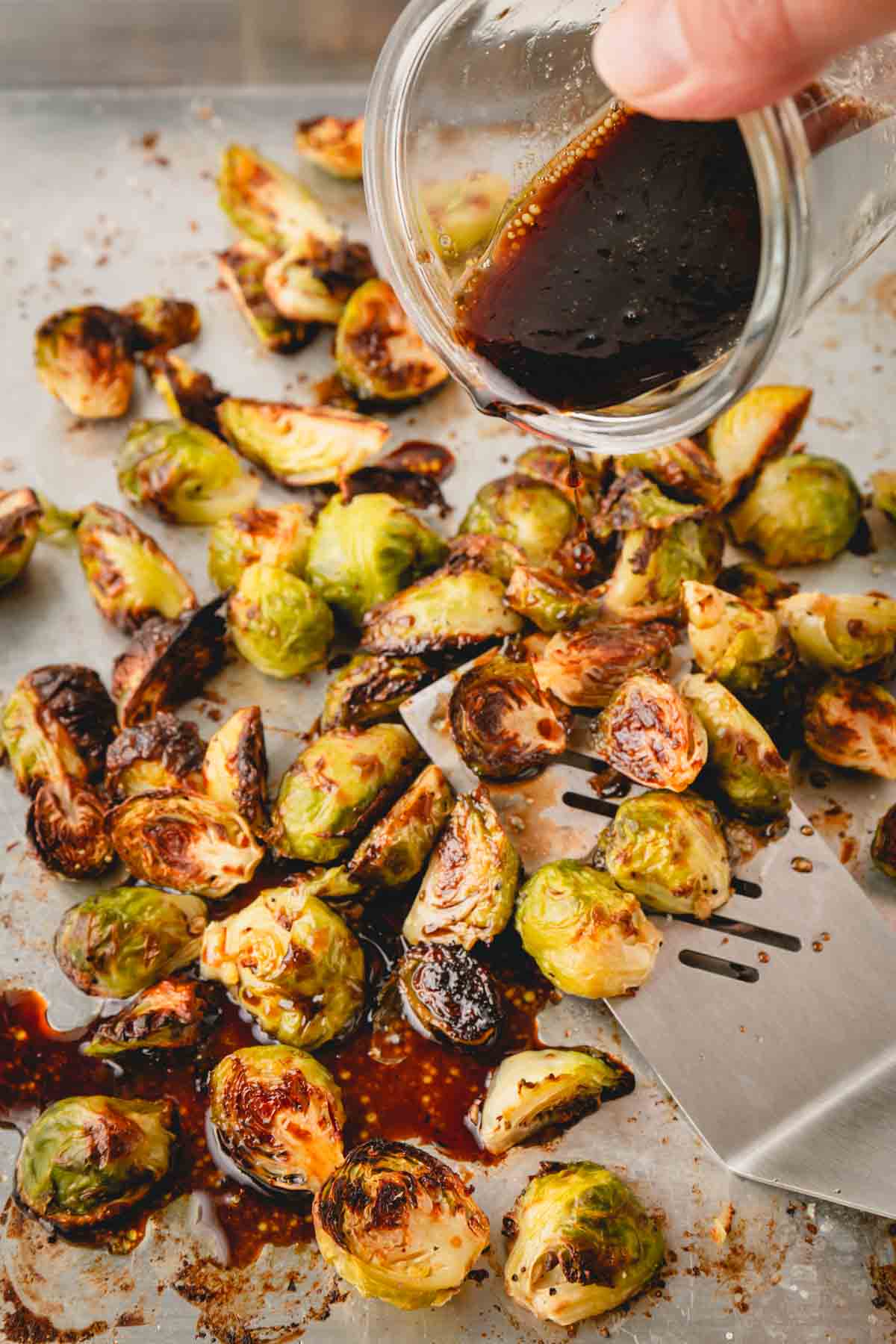 A hand is drizzling the honey balsamic sauce over the roasted Brussels sprouts.