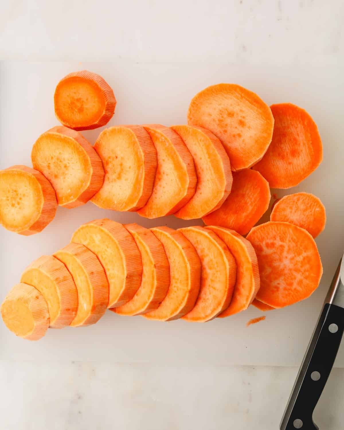 A knife slicing sweet potatoes into rounds on a cutting board.