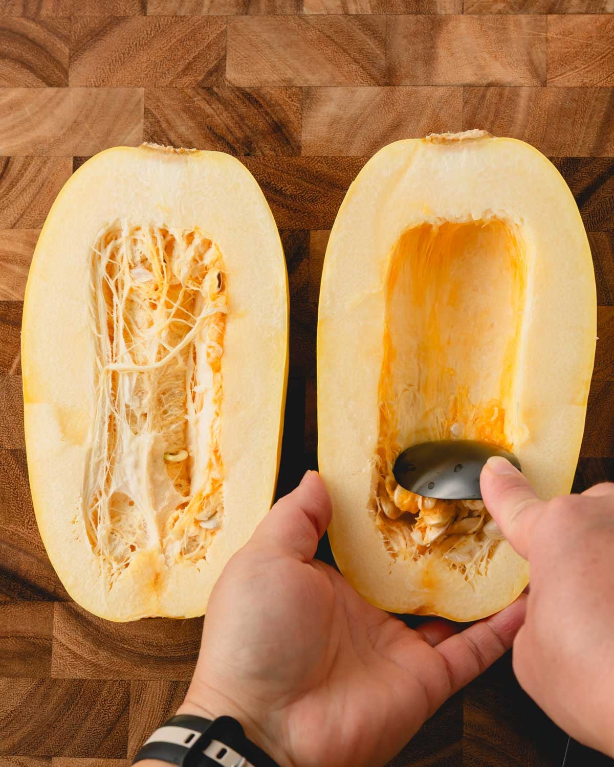 Two hands using a black spoon to scrape out the seeds of the spaghetti squash.