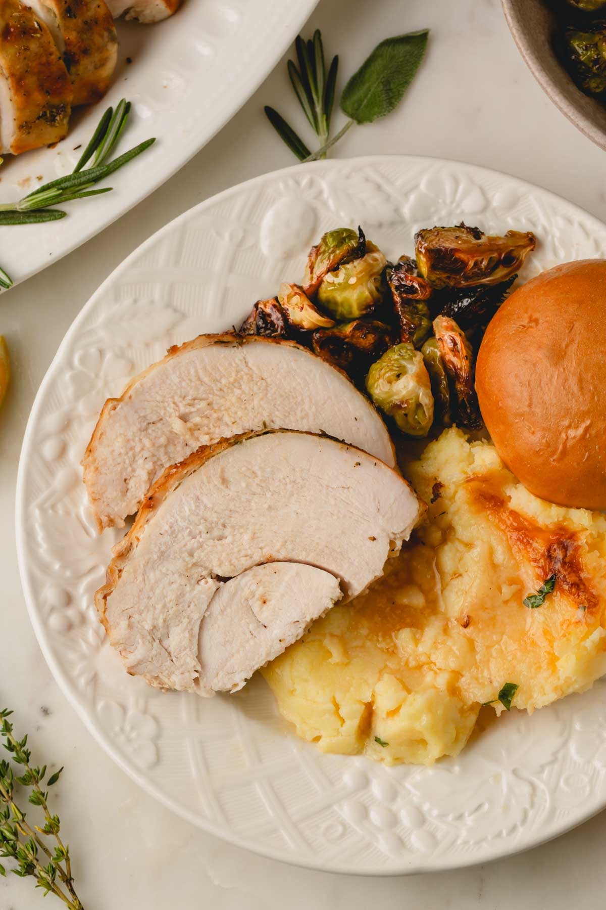 A white plate with a couple of slices of turkey breast, Brussels sprouts, mashed potatoes, and a roll.