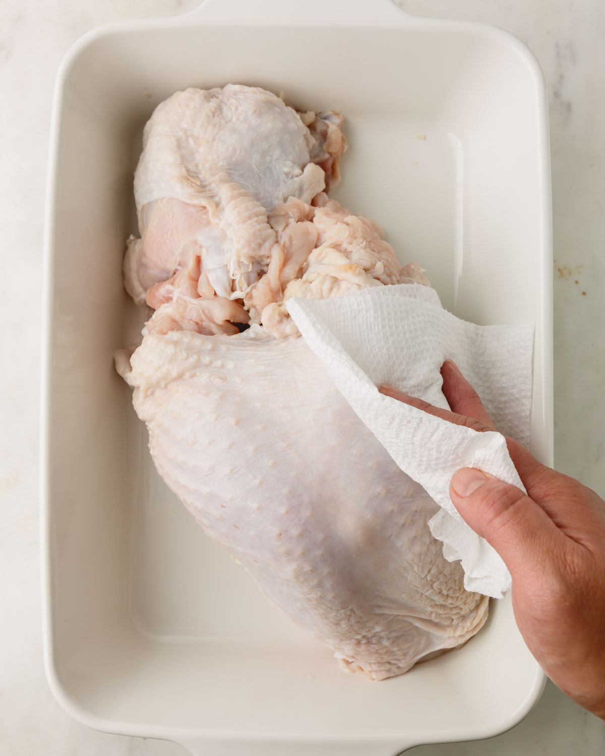 A white baking dish with turkey breast. A hand is patting the skin dry with paper towels.