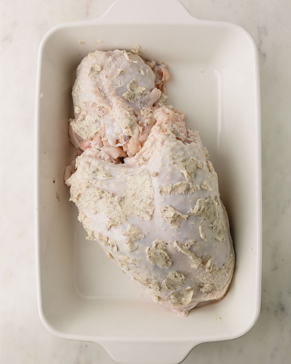 A white baking dish with a turkey breast that has herb butter rubbed in it.
