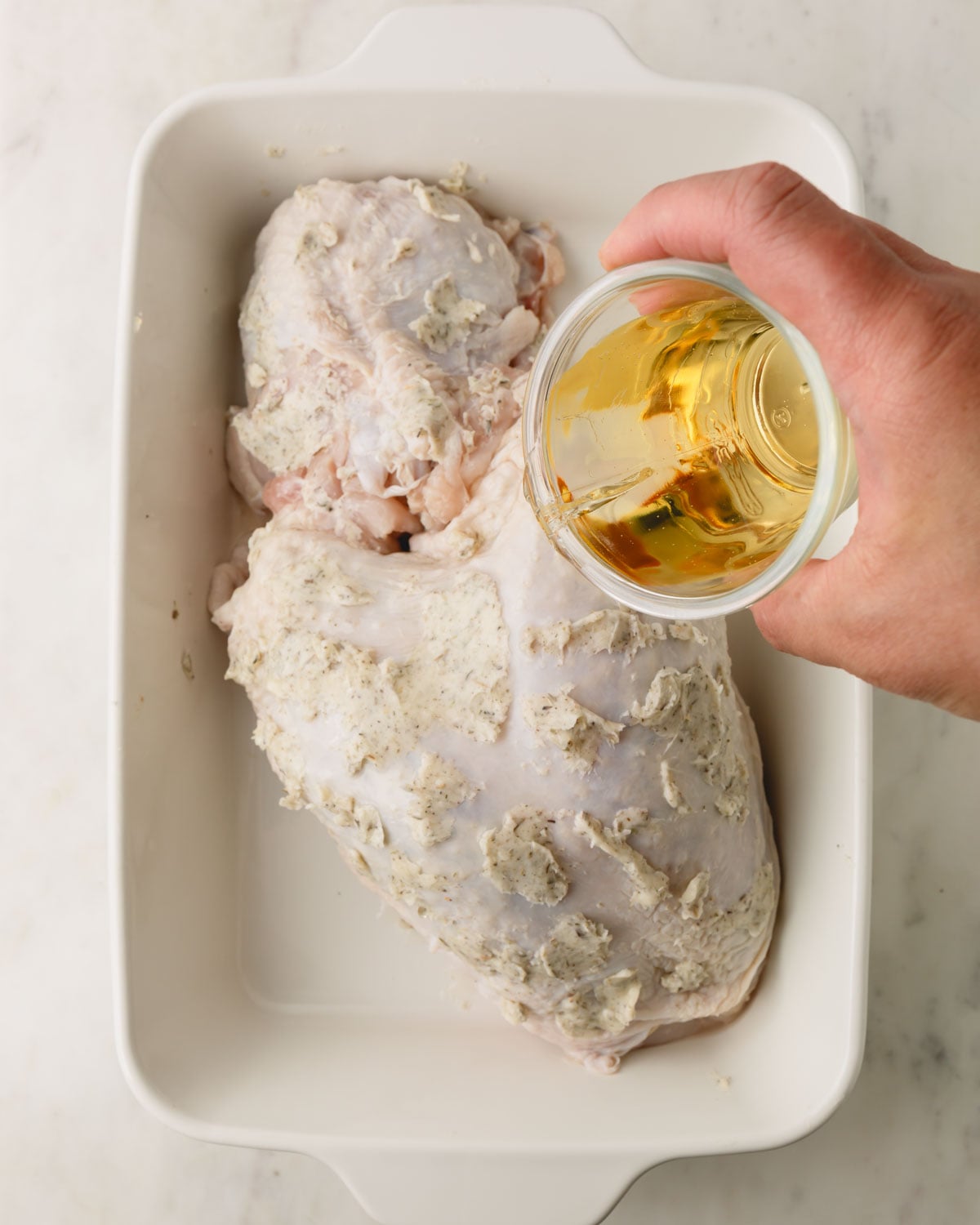 A hand is pouring a cup of white wine cover the turkey breast in a white baking dish.