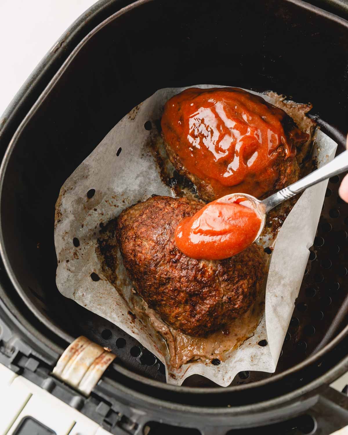 Meatloaf in the air fryer basket with a spoon spreading ketchup glaze on top.