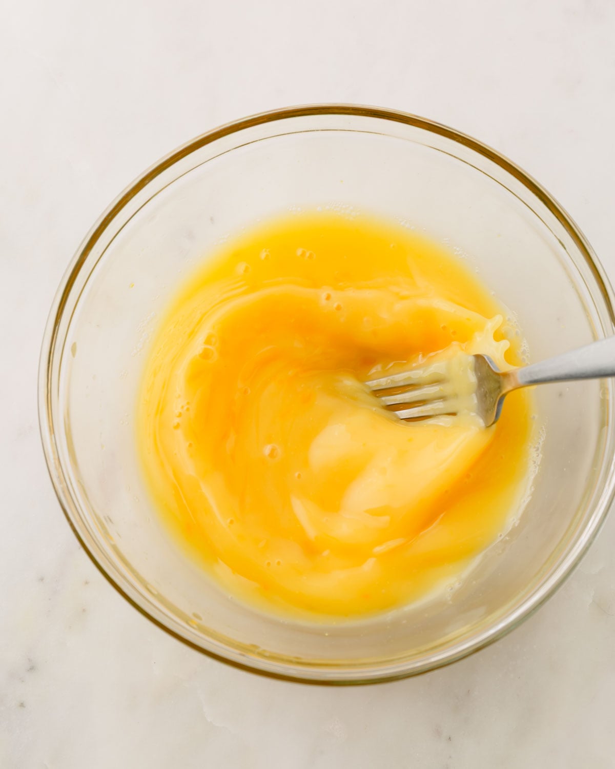 A glass bowl with a fork mixing eggs and chicken broth together.