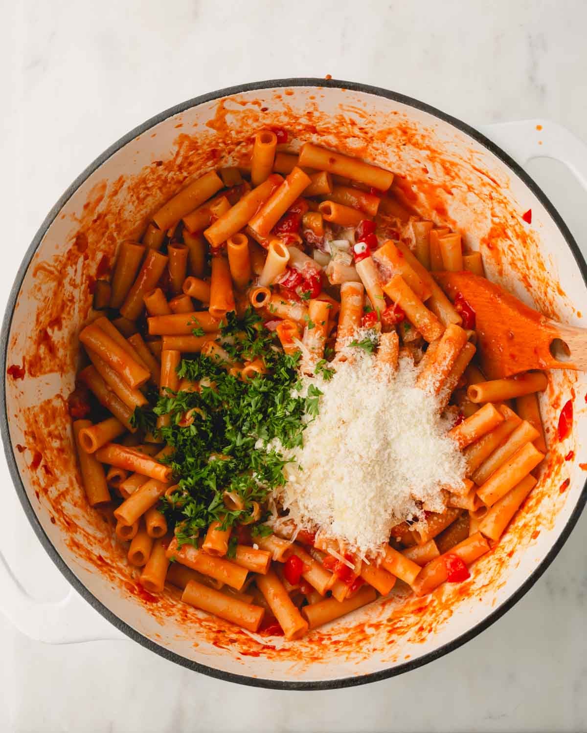 Cooked ziti noodles in marinara sauce with basil, cream, and parmesan cheese and a wooden spoon in a white skillet.