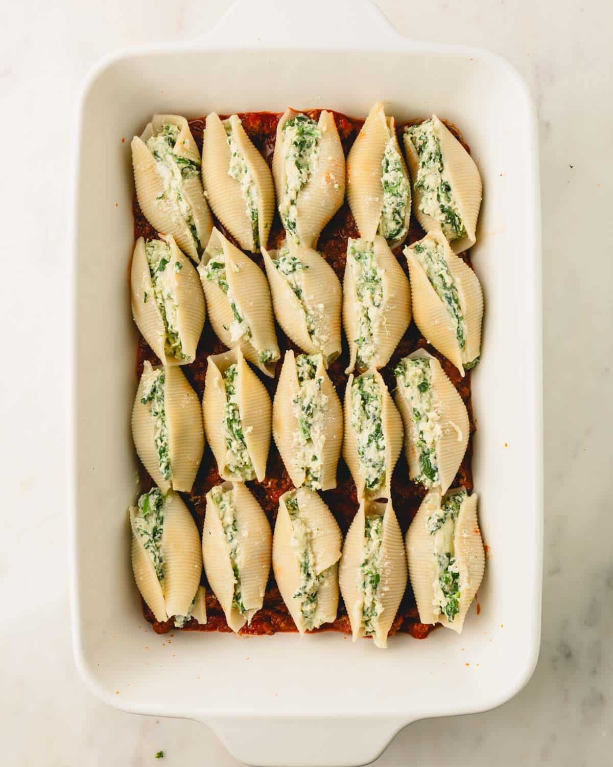 A white casserole dish with stuffed shells lined up on top of the meat sauce.