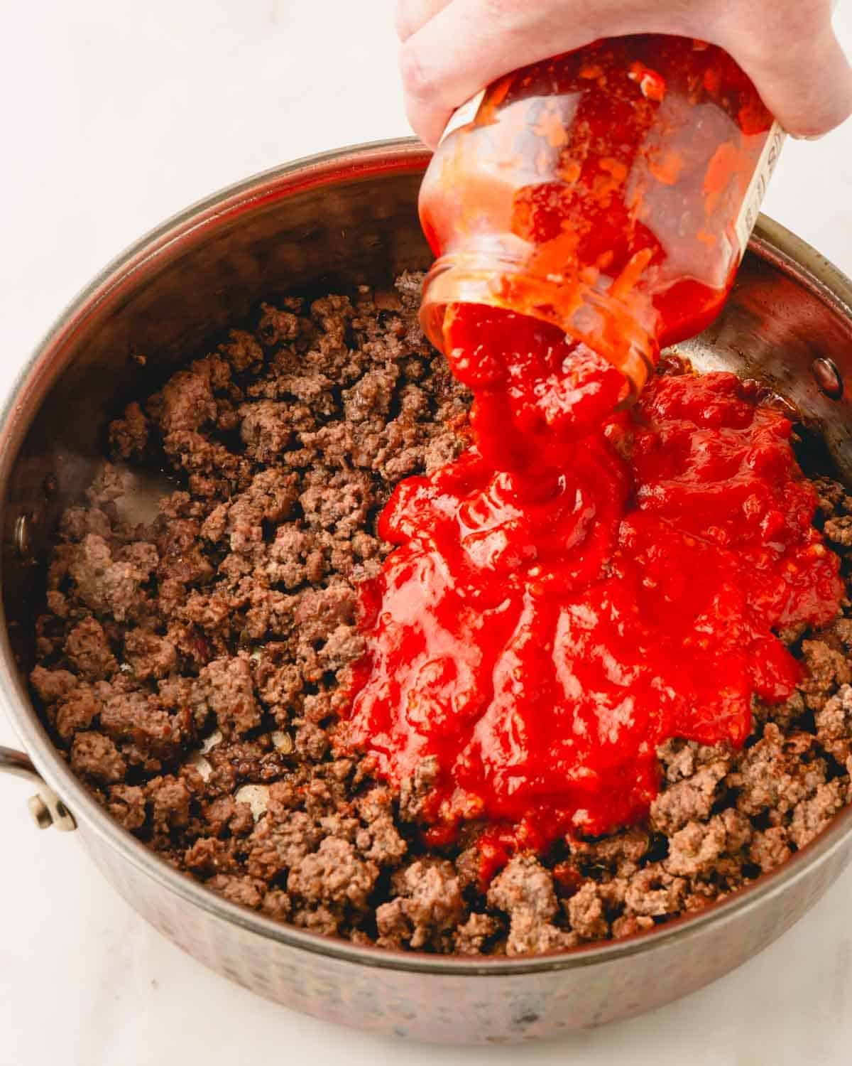 Jarred tomato sauce is being added to a skillet with ground beef and spices.