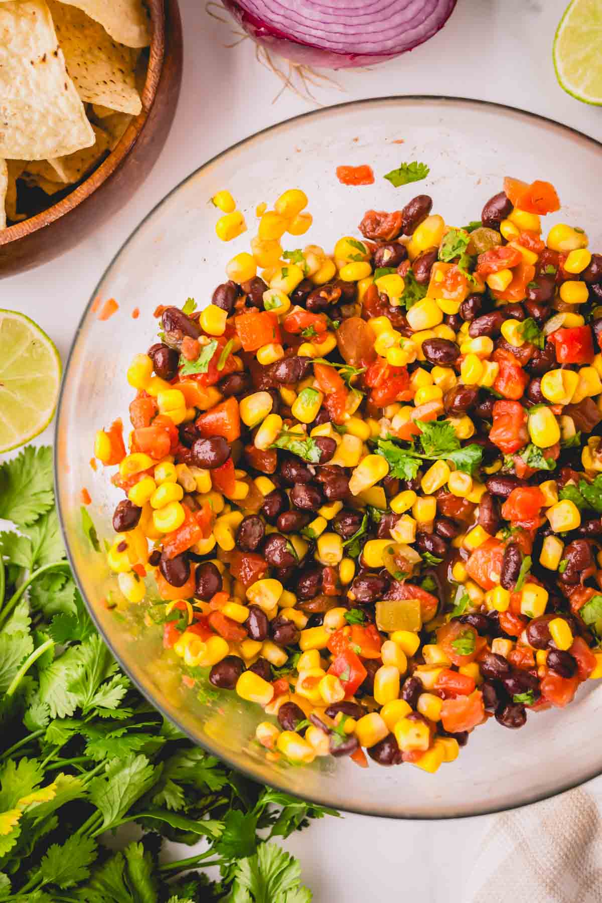 A glass bowl with corn and black bean salsa with cilantro and chip in the background.