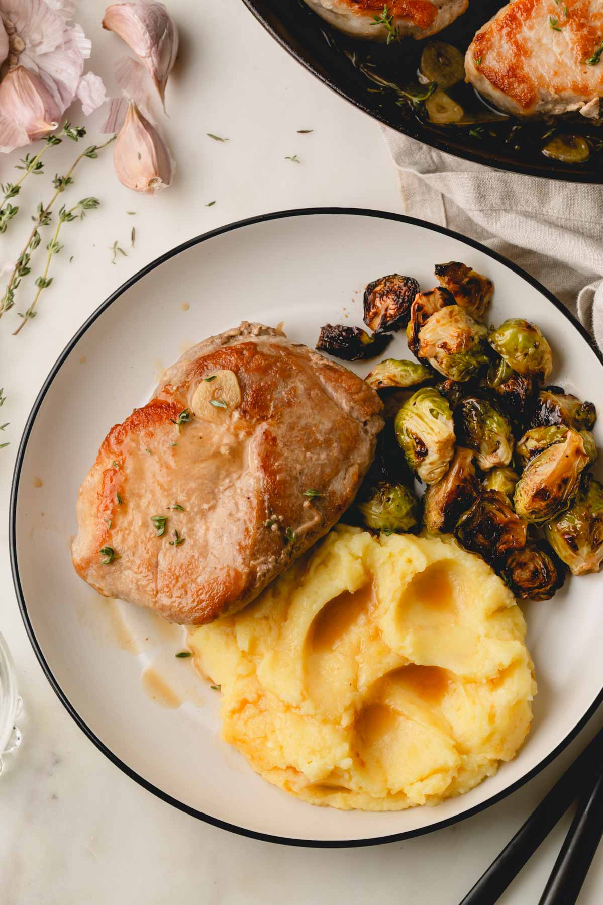 A white plate with a cast iron pork chop, mashed potatoes, and Brussels sprouts. Garlic cloves and more pork chops in the background.