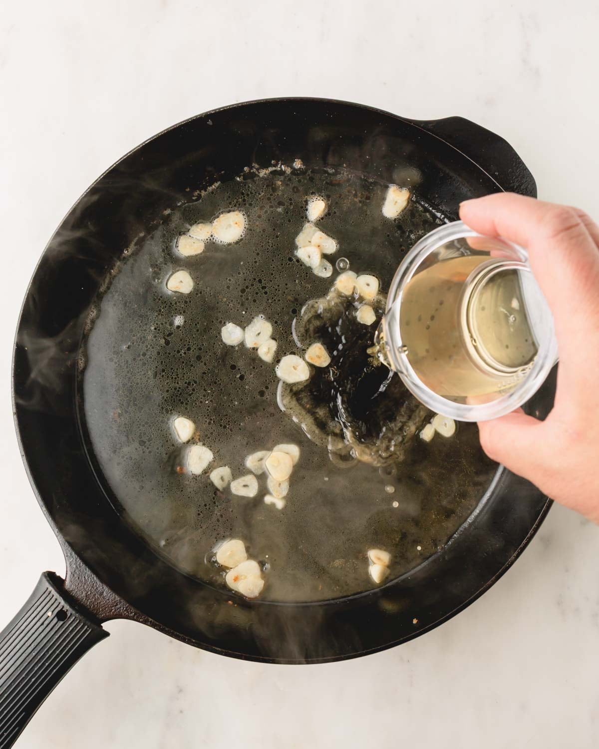 A cast iron skillet with garlic and a hand adding white wine from a bowl.