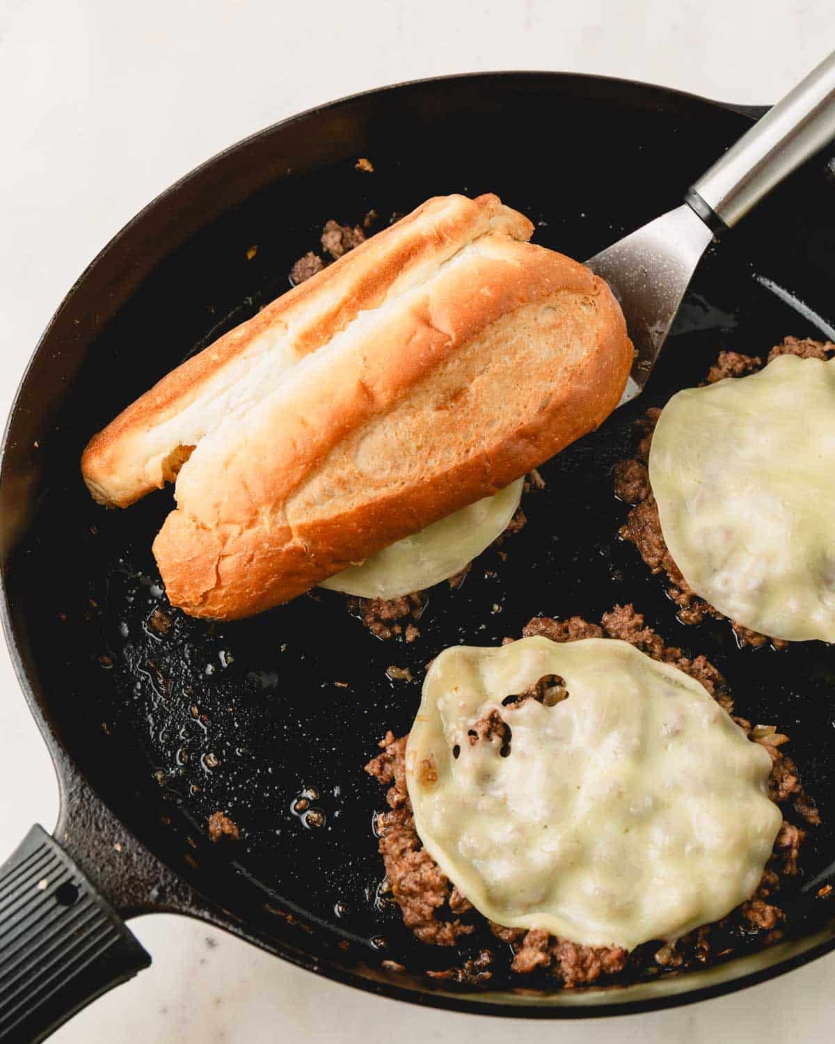 A bun is placed over the pile of ground beef and melted cheese in the skillet with a stainless steel spatula underneath.