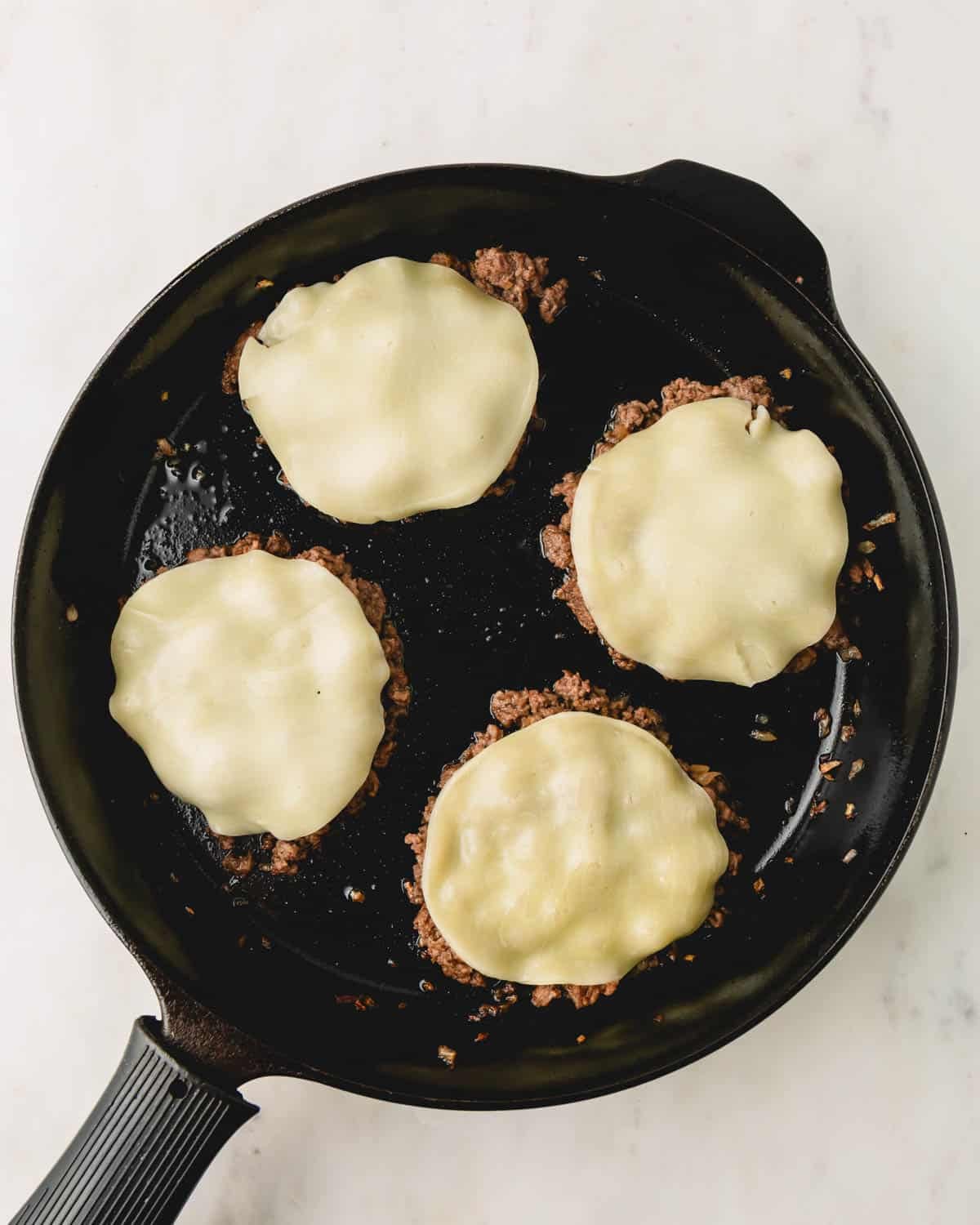 Slices of provolone cheese melting on top of four piles of ground beef in a skillet.