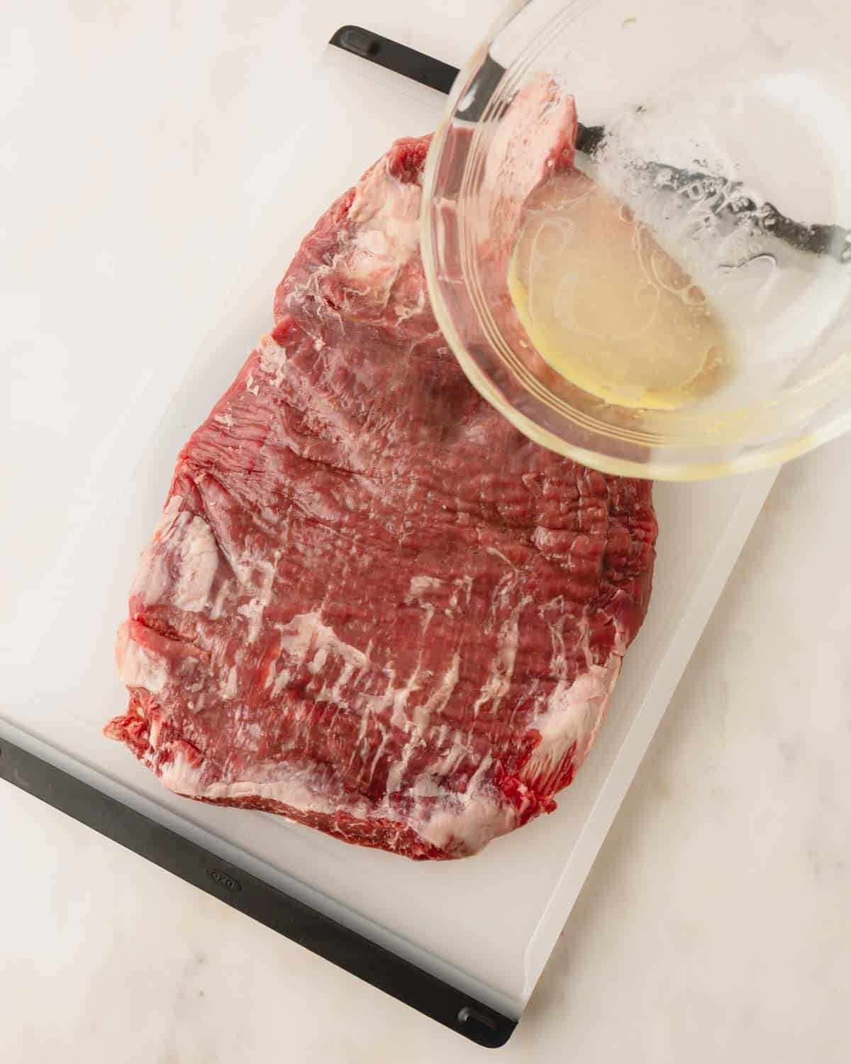Flank steak on a plastic cutting boards with the lime marinade being poured over from a glass bowl.