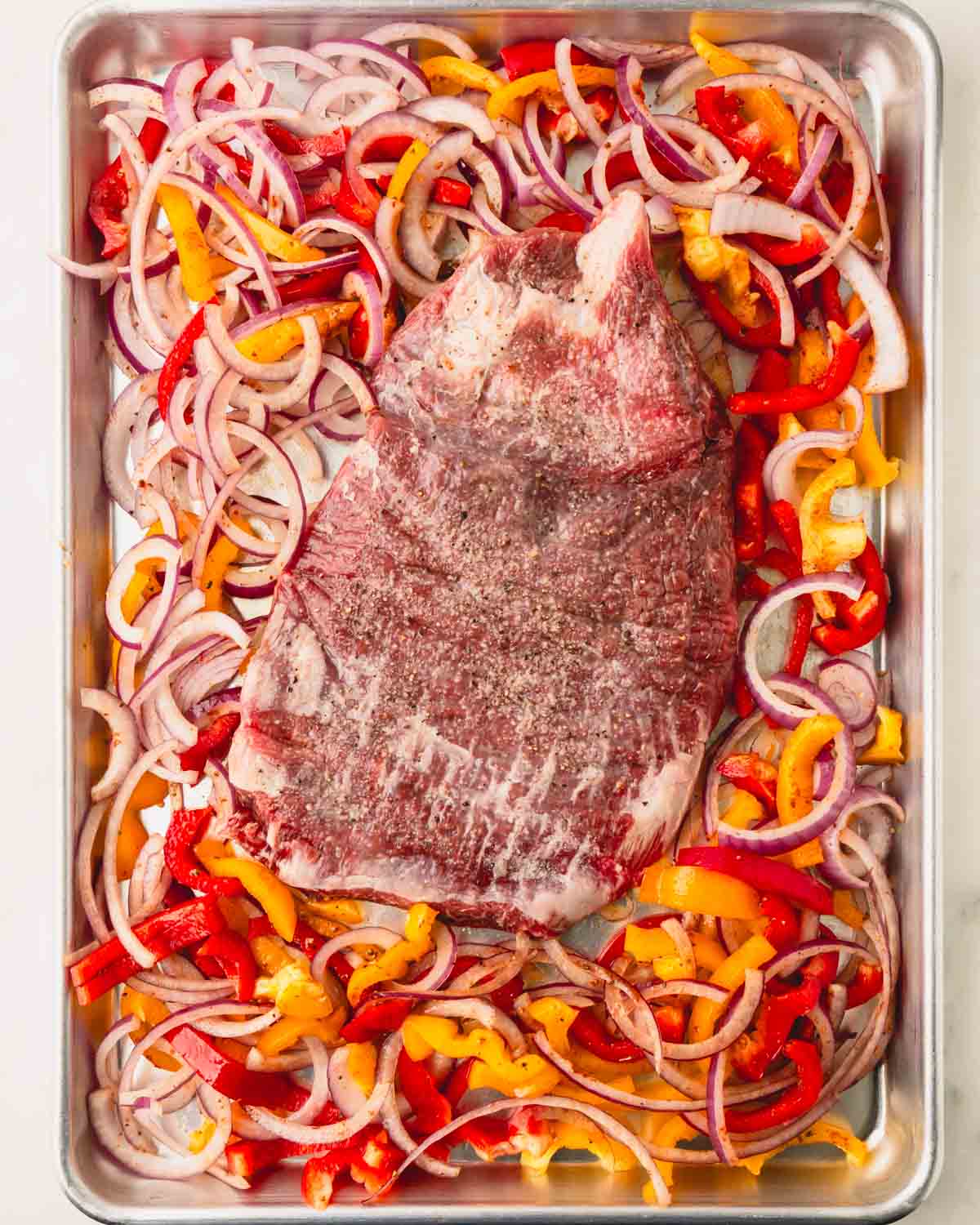 A sheet pan with sliced onions, bell peppers, and flank steak with seasoning.