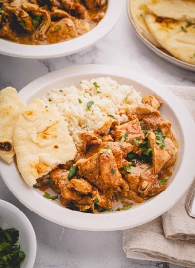 Slow cooker butter chicken plated with rice and naan on a white plate with more butter chicken and naan in the background.