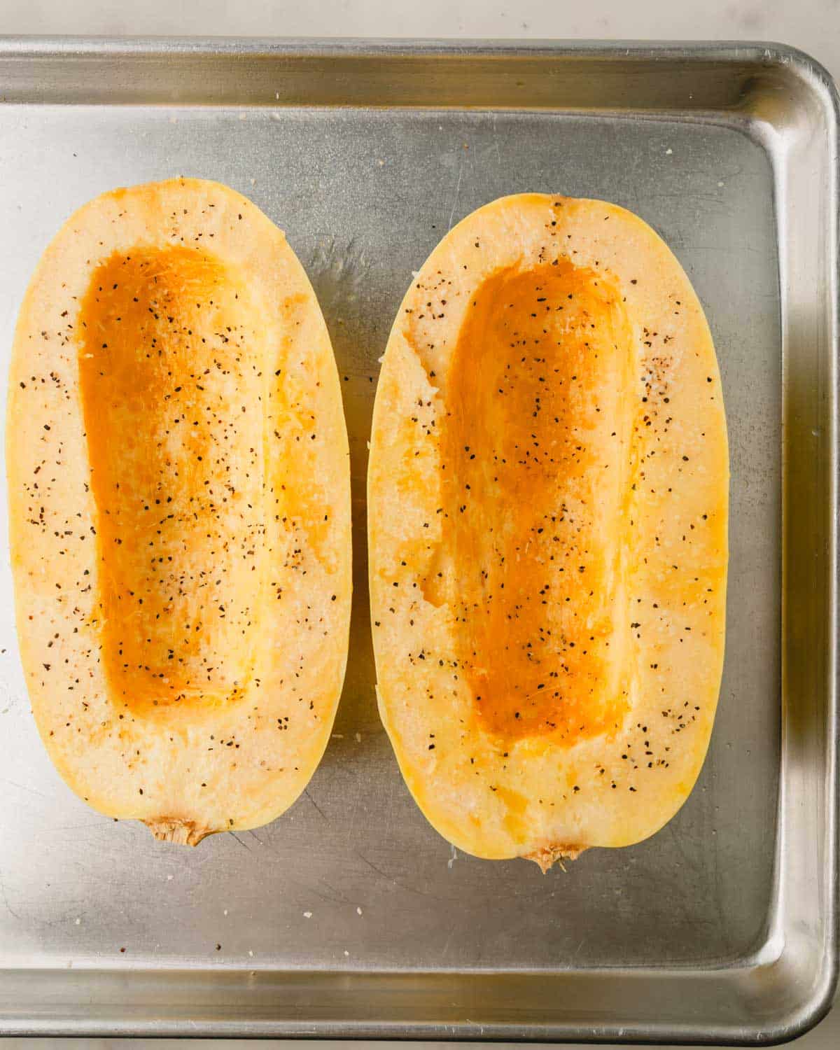 Two halves of spaghetti squash are on a baking sheet with salt and pepper.