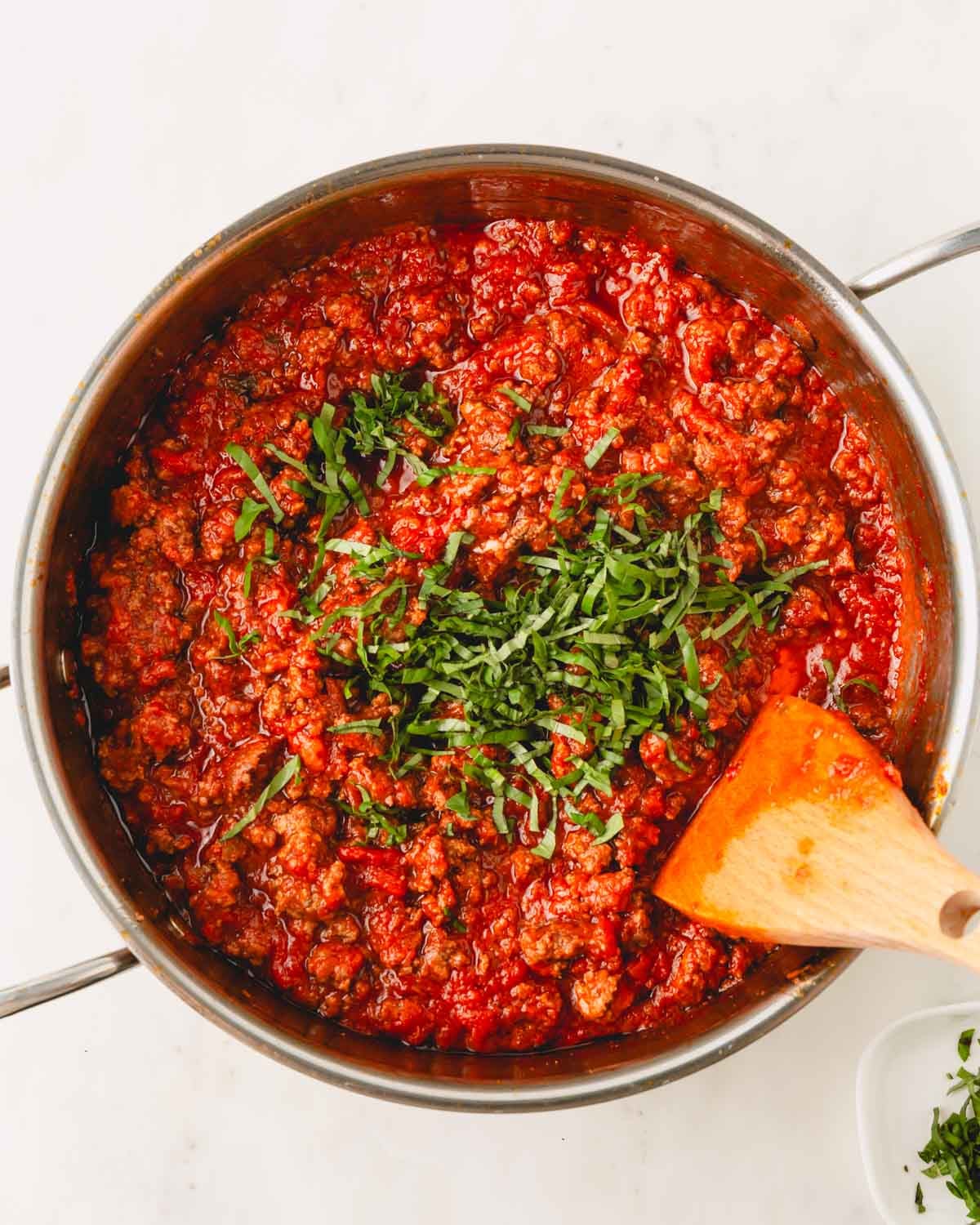 A skillet with ground beef tomato sauce, chopped basil and a wooden spoon.
