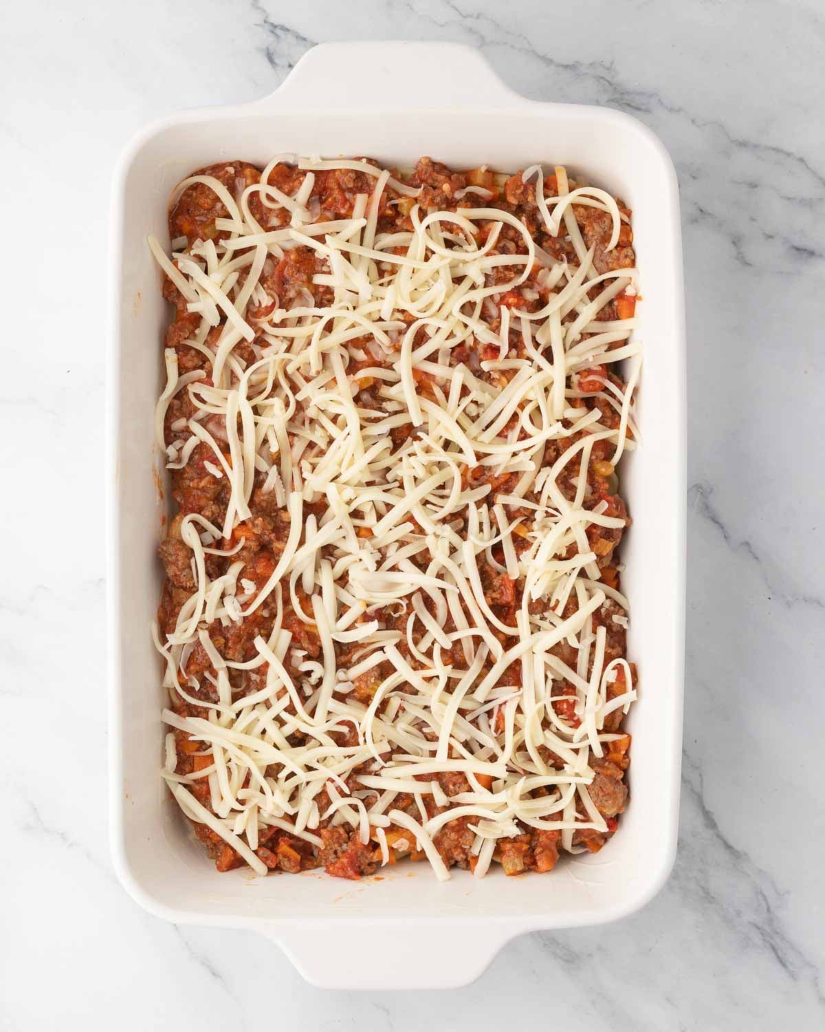 A white baking dish with penne noodles, meat sauce, and shredded mozzarella cheese on top.