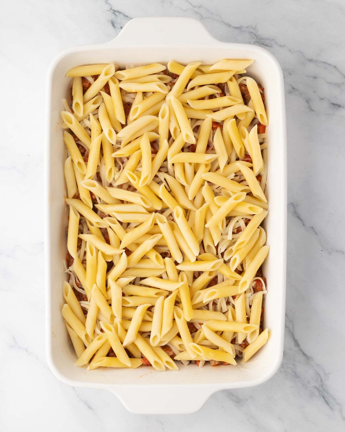 A white baking dish with meat sauce, mozzarella cheese, and a second layer of boiled penne noodles on top.