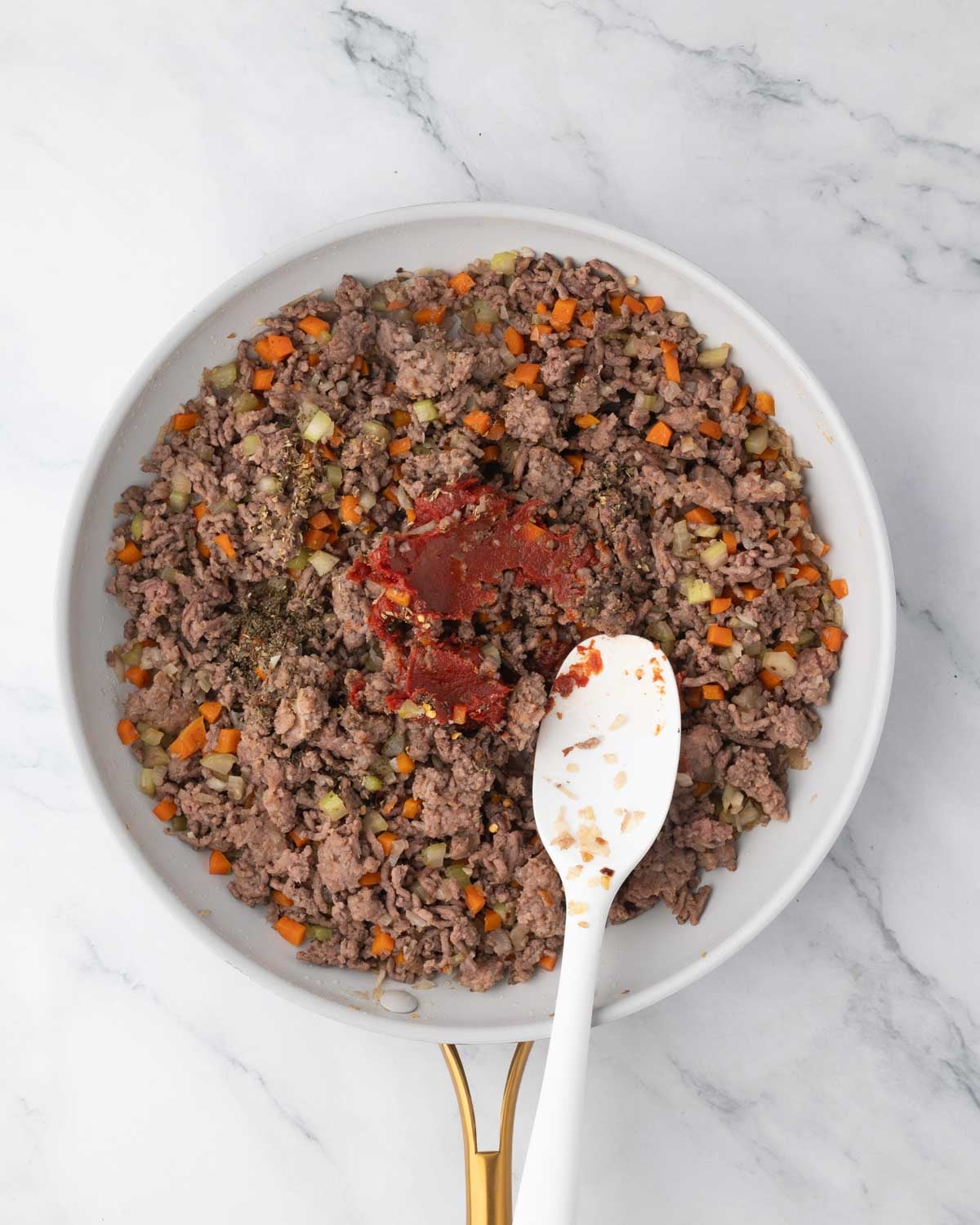 A white skillet with ground beef, celery, onions, and carrots cooking with tomato paste added. A white spatula is in the pan.
