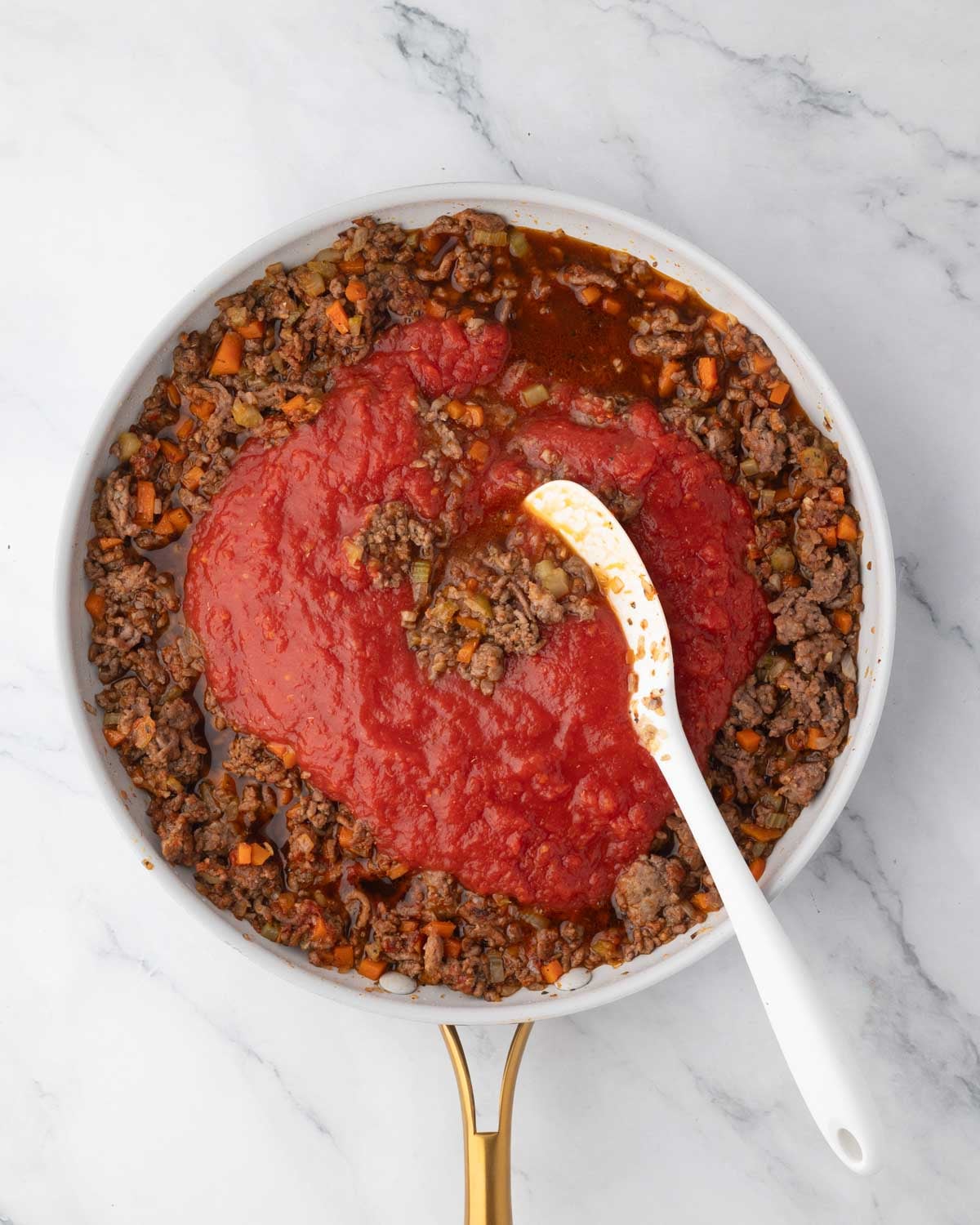 A white skillet with ground beef, veggies, and canned tomatoes cooking with a white spatula.