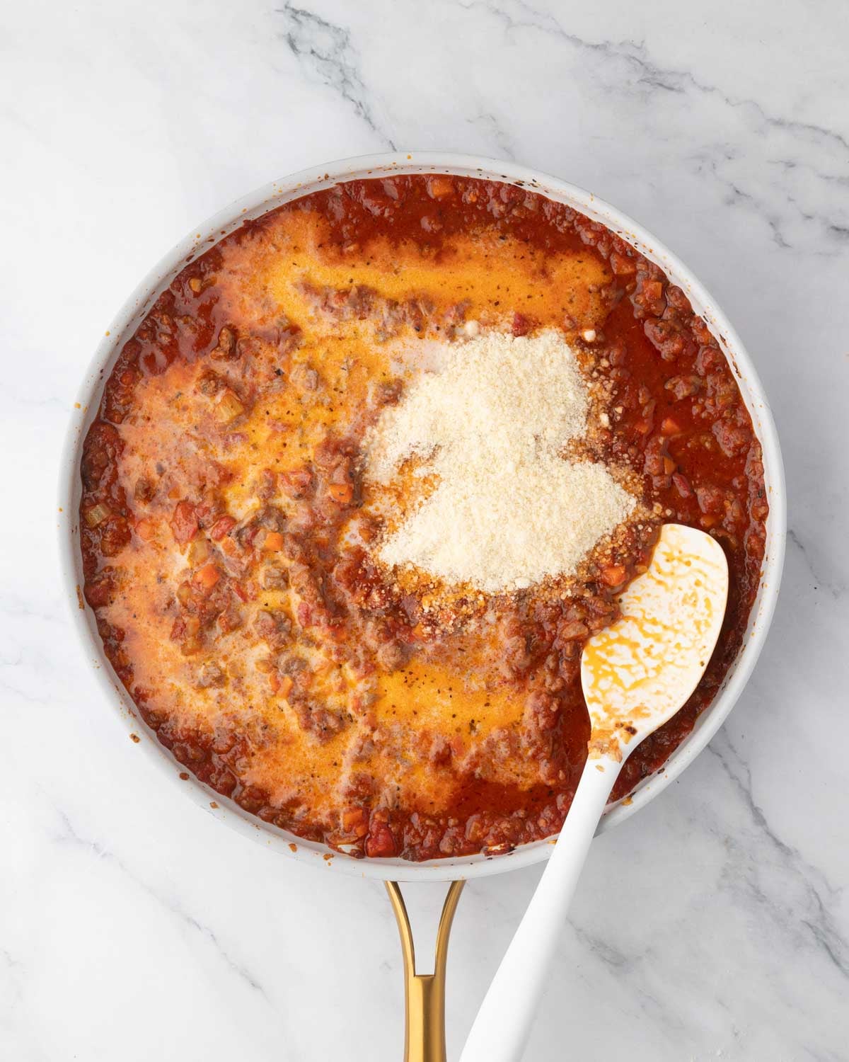 A white skillet with ground beet tomato sauce cooking with parmesan cheese added on top and a white spatula.