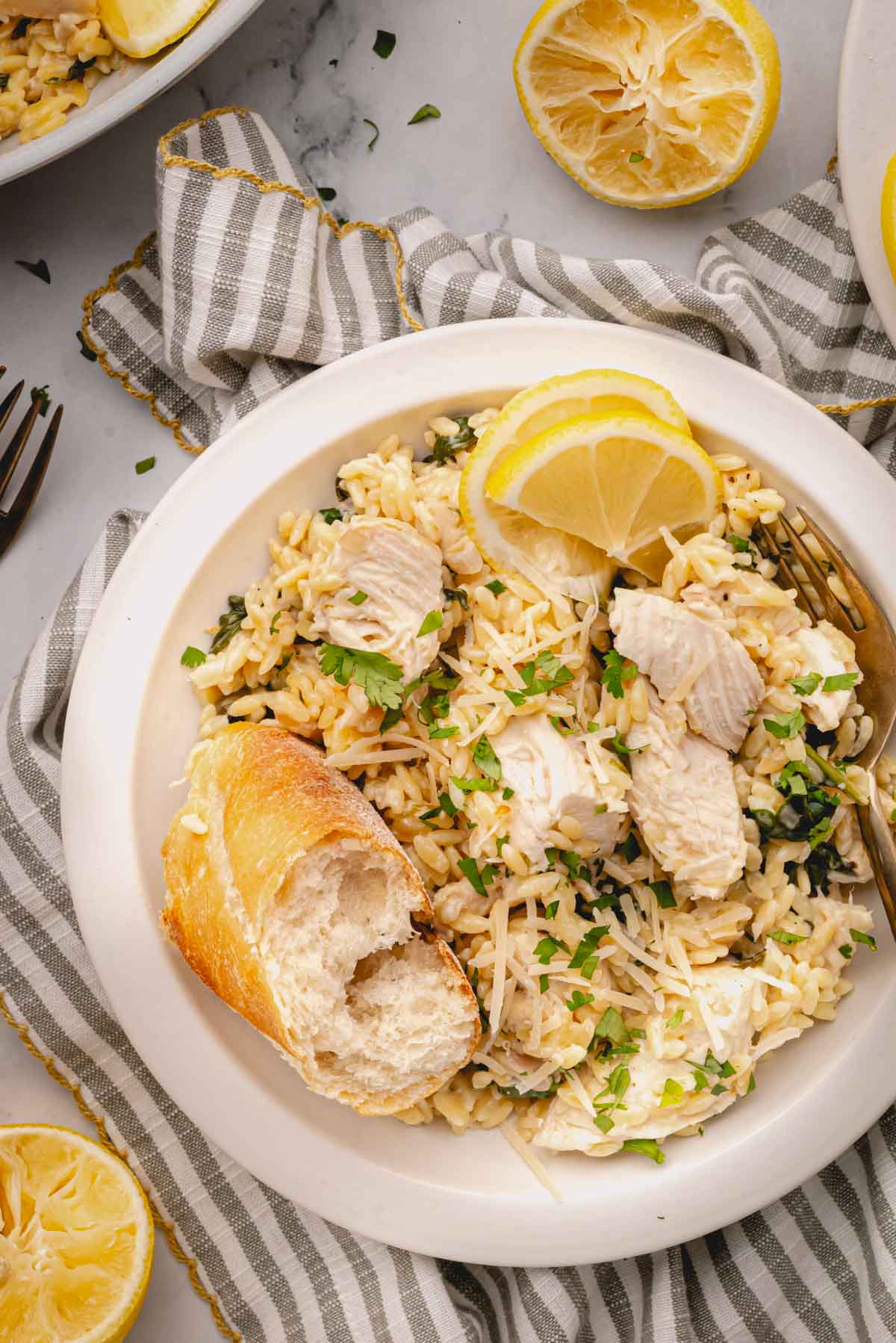 A white bowl of lemon chicken orzo with bread and lemon slices on a striped linen.