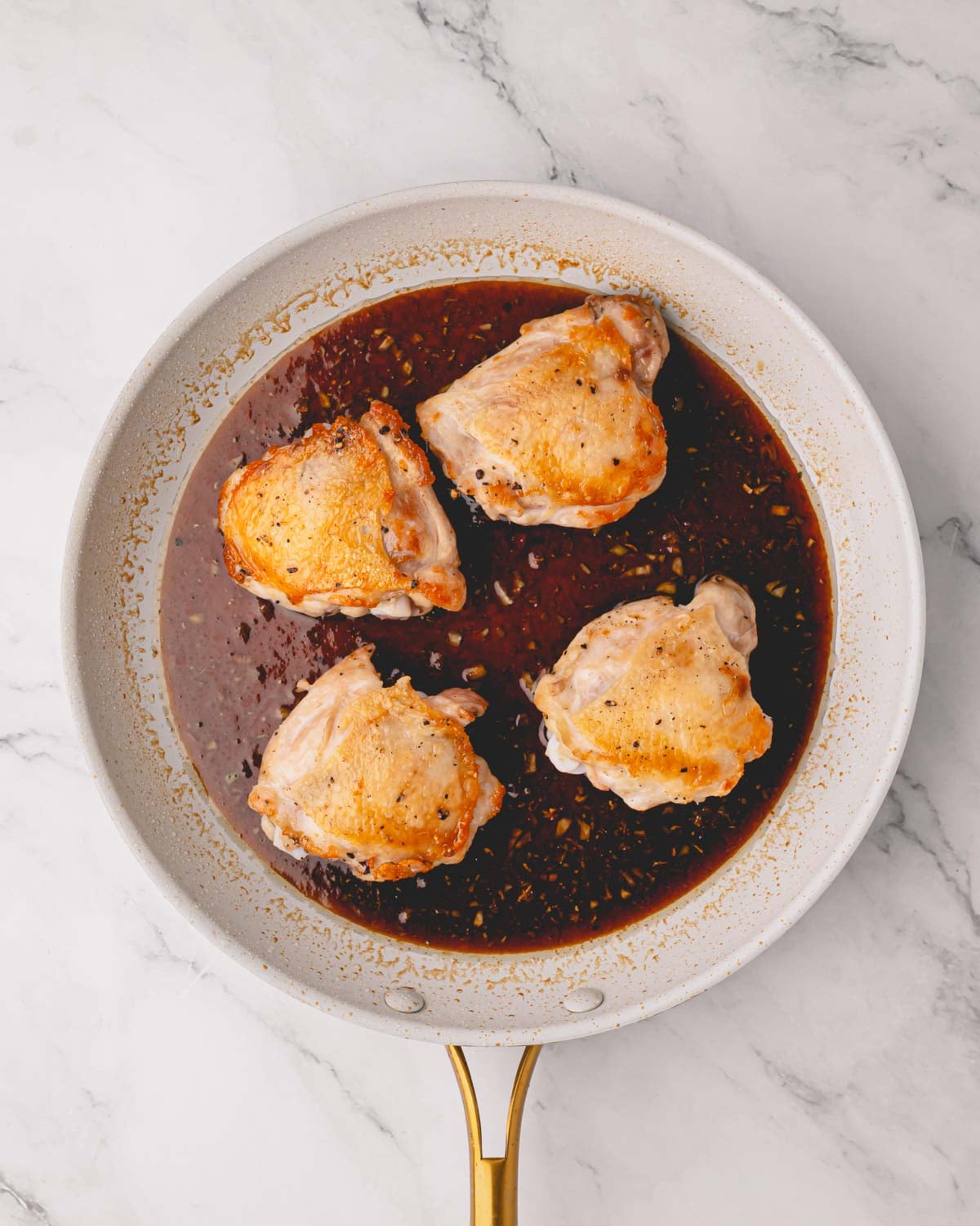 A white skillet with seared chicken thighs and shoyu sauce in the bottom.