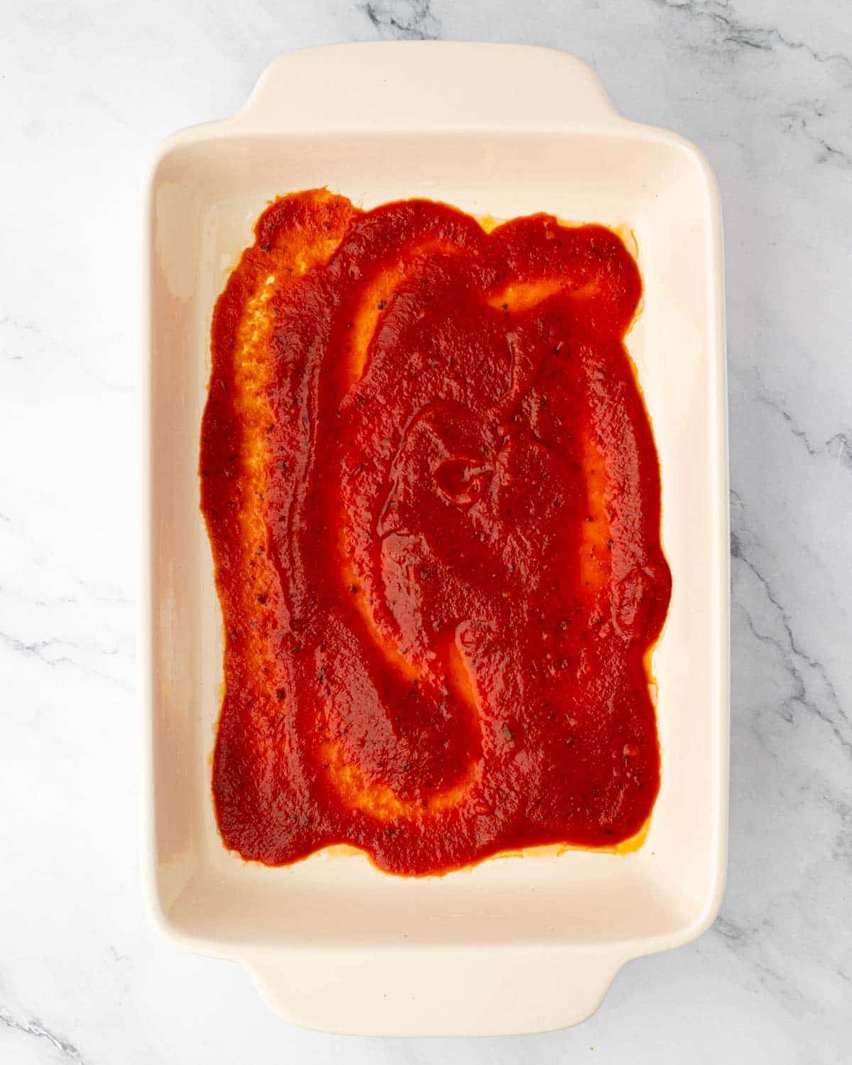 A white baking dish with marinara sauce in the bottom.