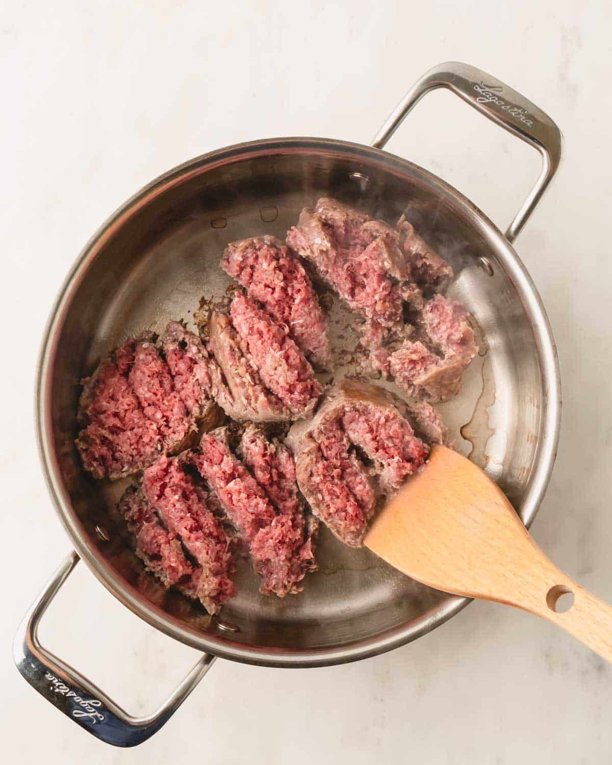 A stainless steel skillet with ground beef browning and a wooden spoon.