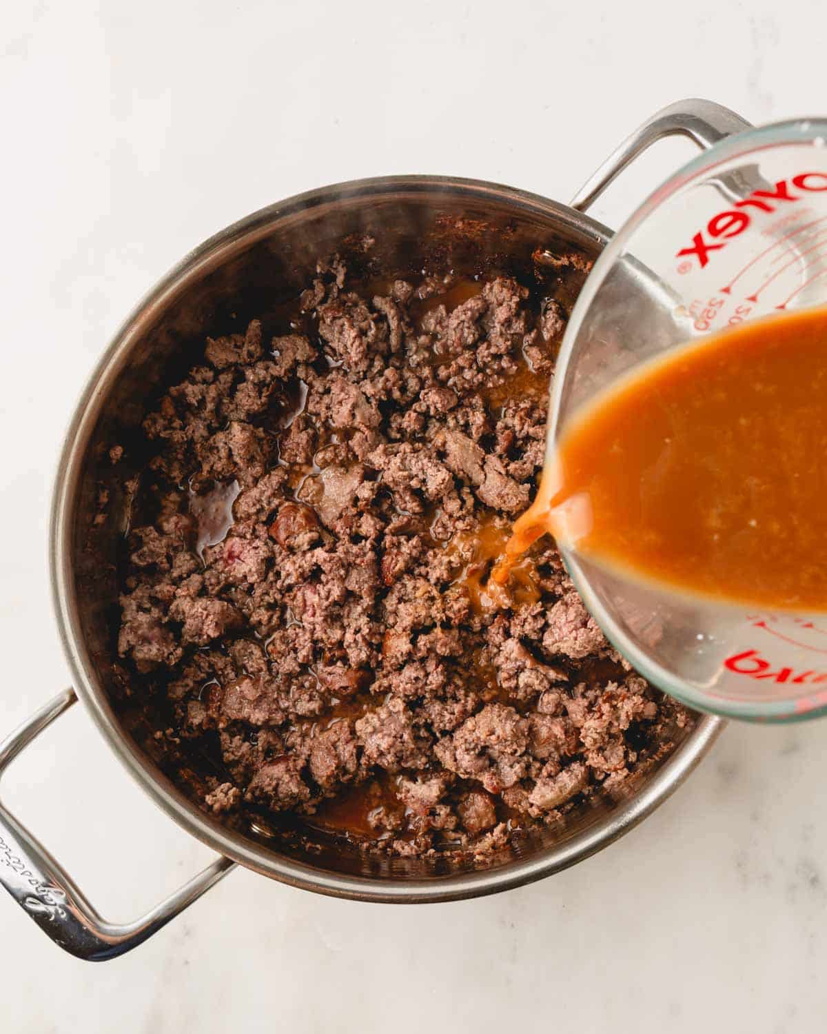 A skillet with ground beef cooking with a measuring cup pouring in the teriyaki sauce.
