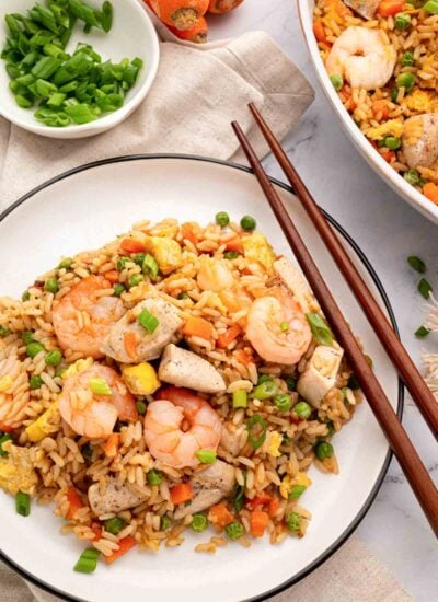 Shrimp and chicken fried rice on a white plate with wooden chopsticks and more fried rice in the background as well as sliced scallions.