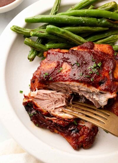 A fork taking a piece of slow cooker BBQ chicken thighs off a white plate. There's a side of green beans on the plate too.