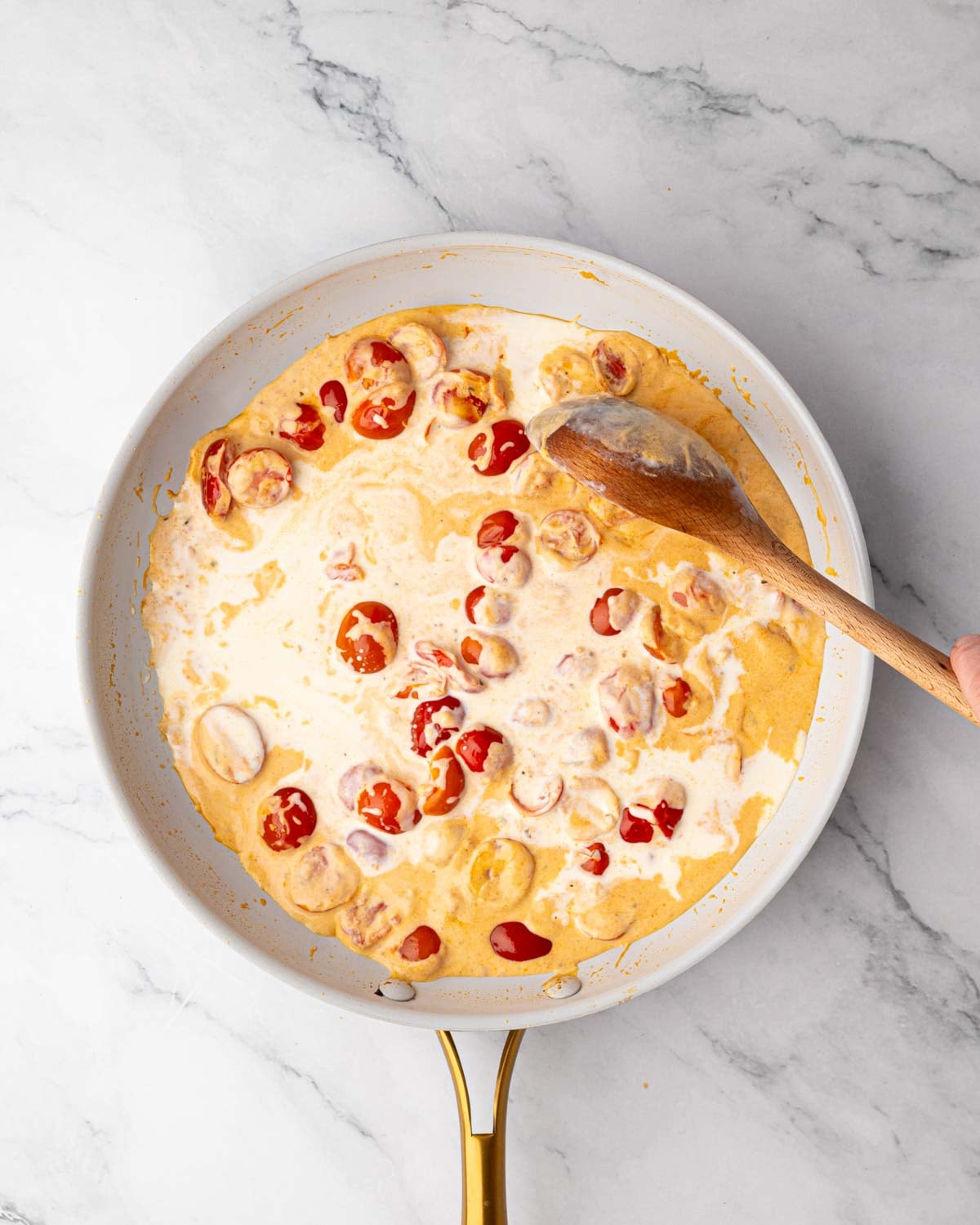 A skillet with a wooden spoon stirring tomato cream sauce.
