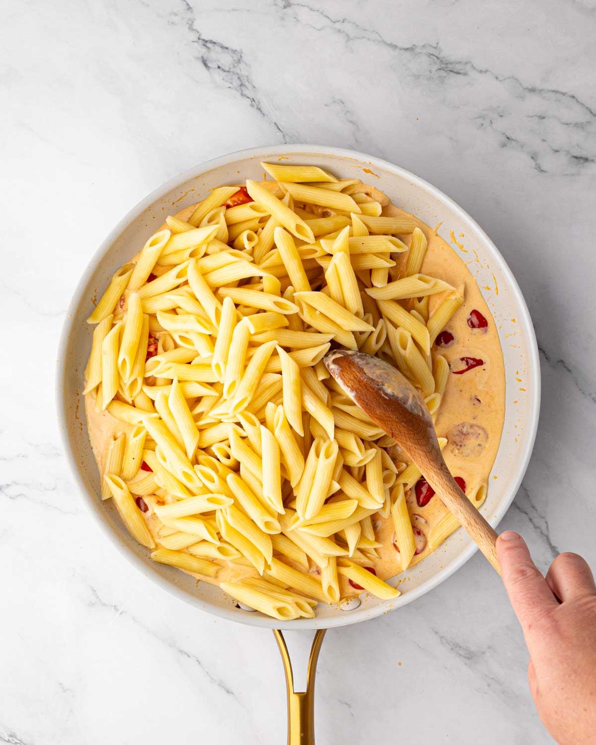 A white skillet with tomato cream sauce and penne pasta being added.