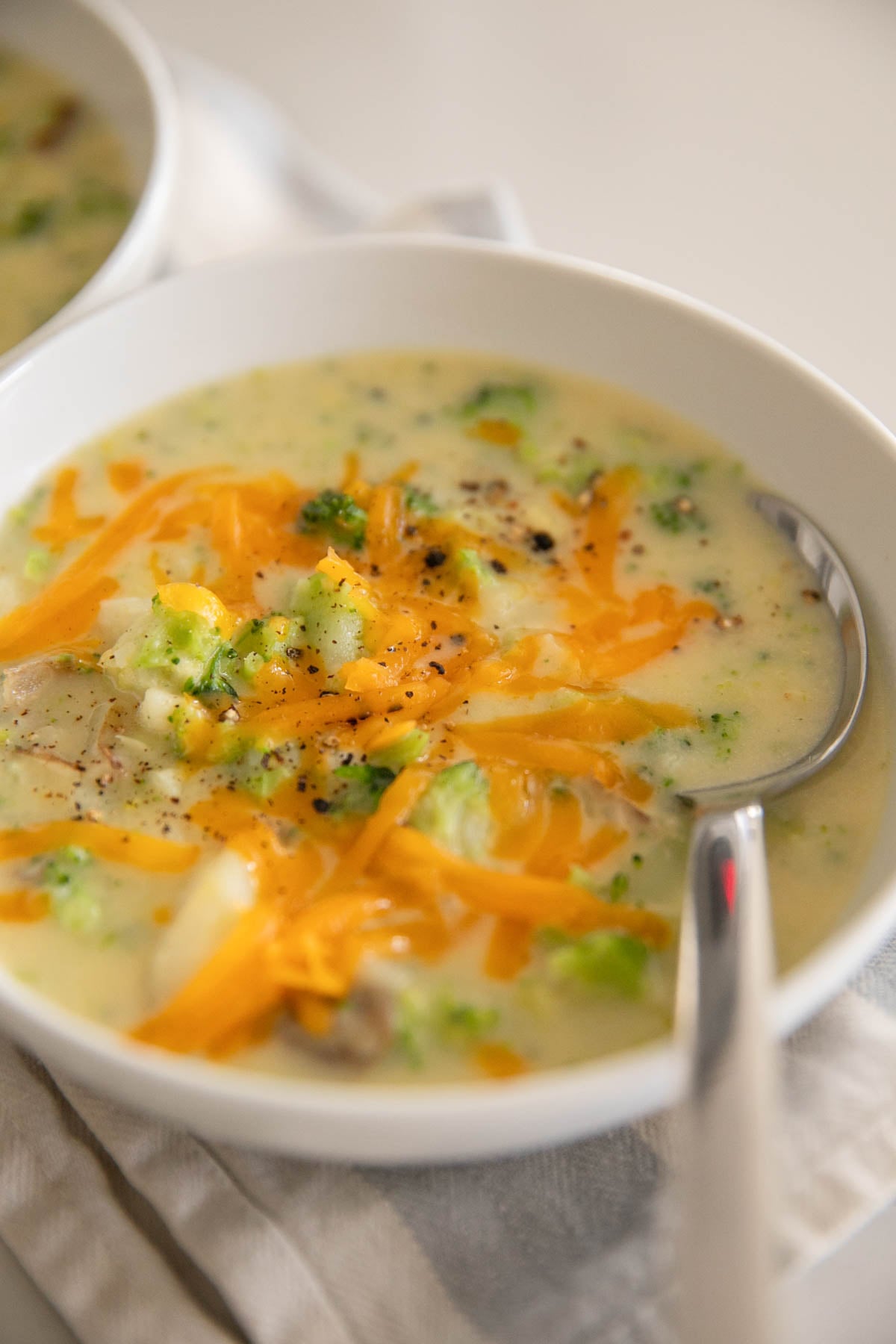 A white bowl with broccoli, cheddar, potato soup and a spoon on the side.