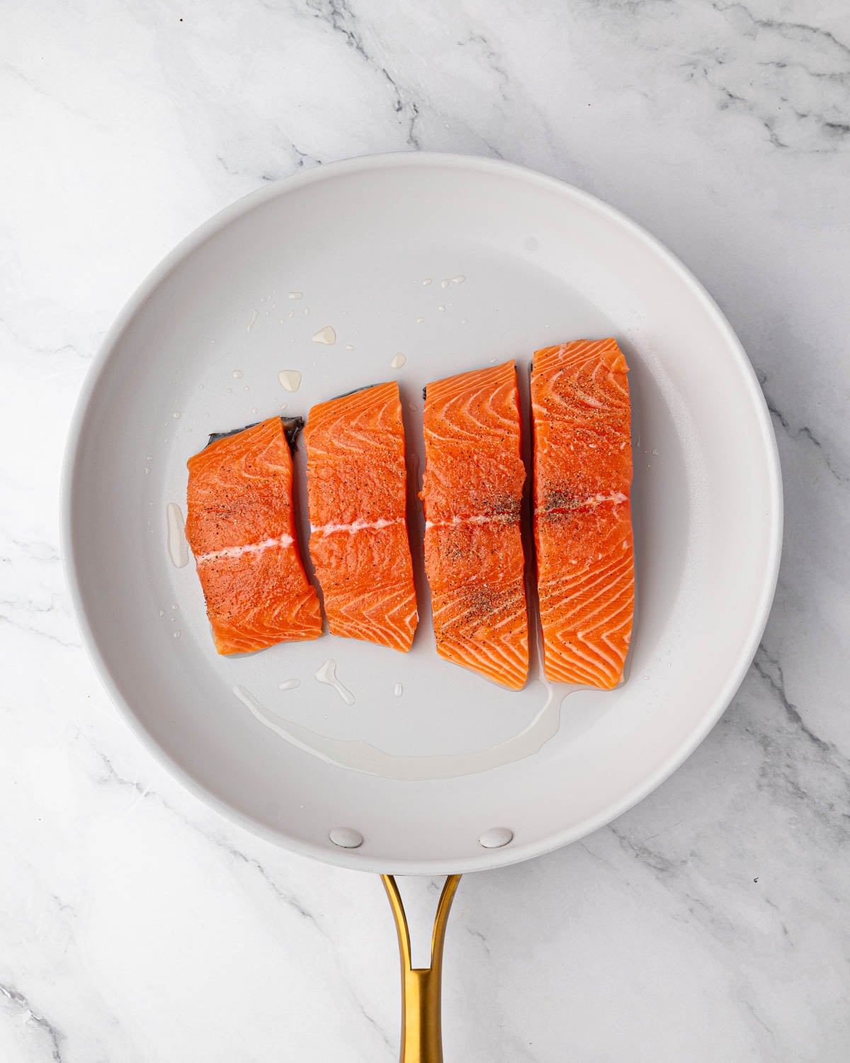 A white skillet with salmon fillets searing.