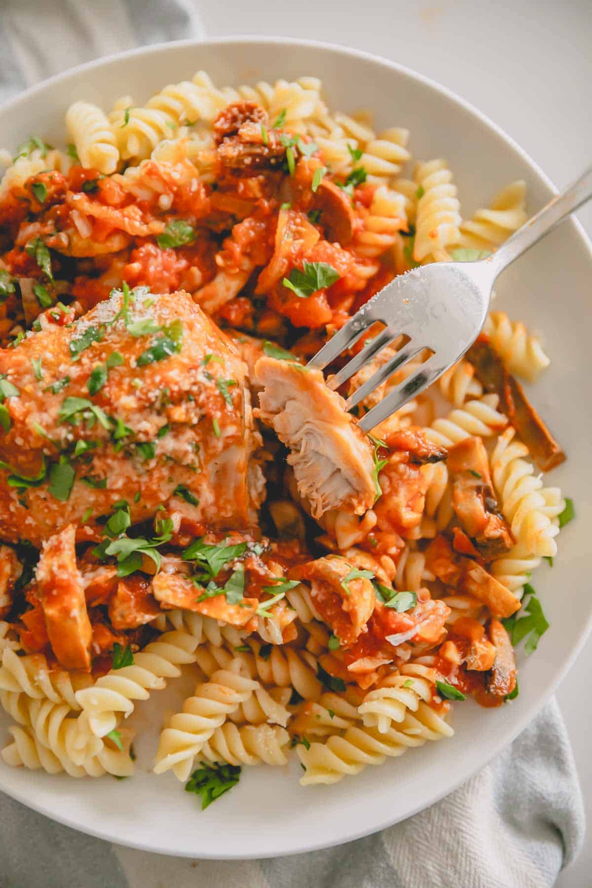 A white plate with chicken cacciatore over pasta with a fork taking a piece of chicken.