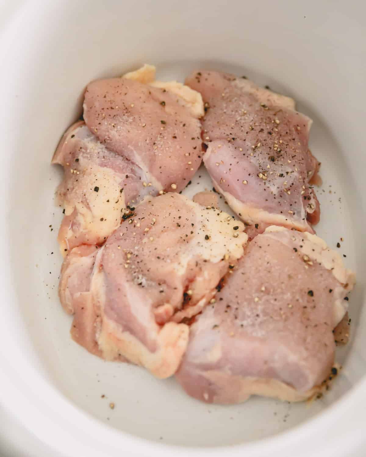 A slow cooker bowl with chicken thighs seasoned with salt and pepper.