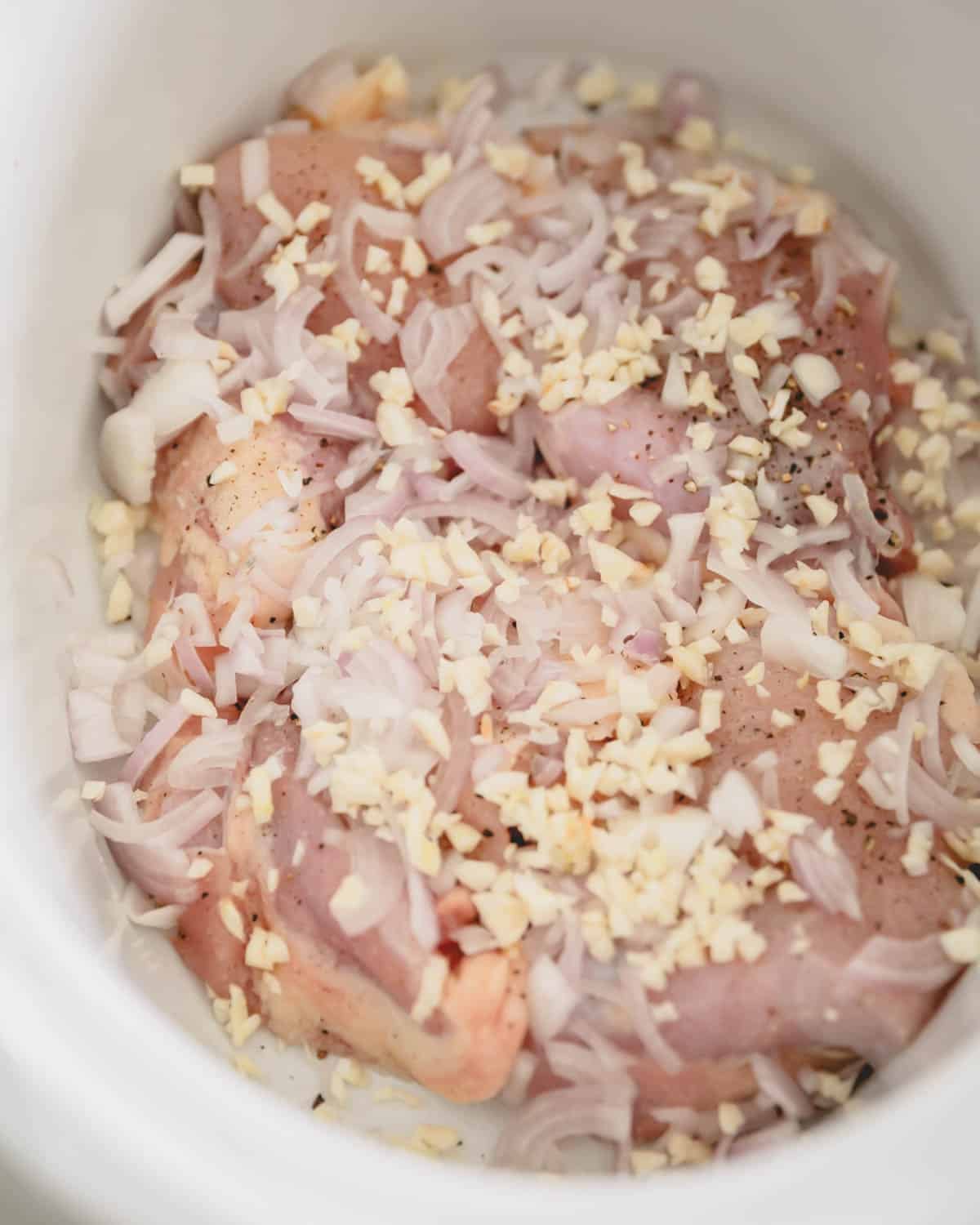 A slow cooker with chicken thighs, shallot, and garlic.