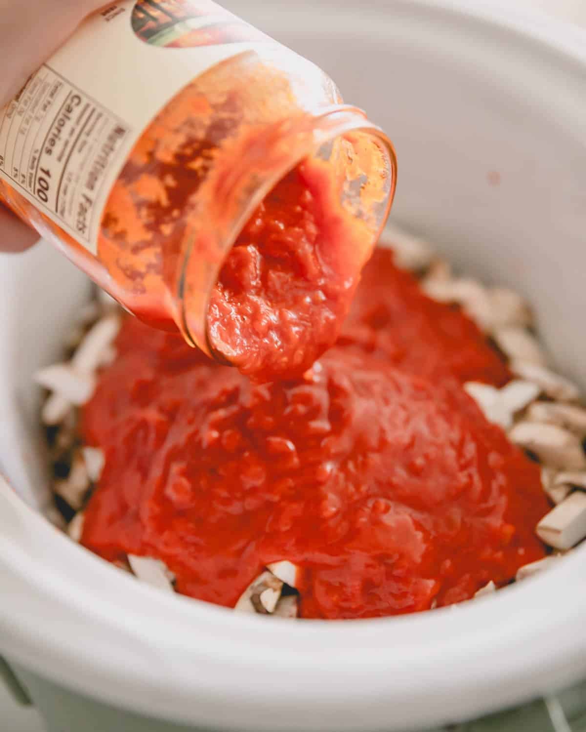 A hand pouring in jarred marinara sauce into a slow cooker for chicken cacciatore.