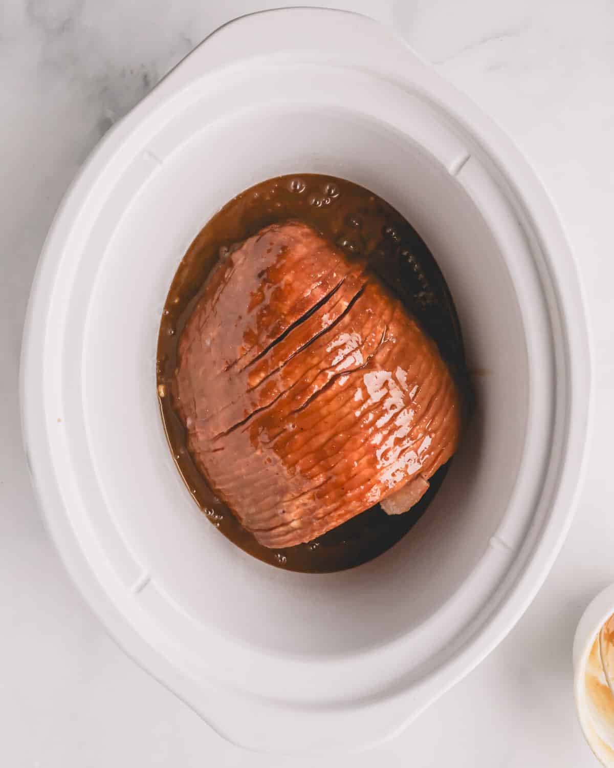 A white crock pot with a ham and honey glaze in the bottom.
