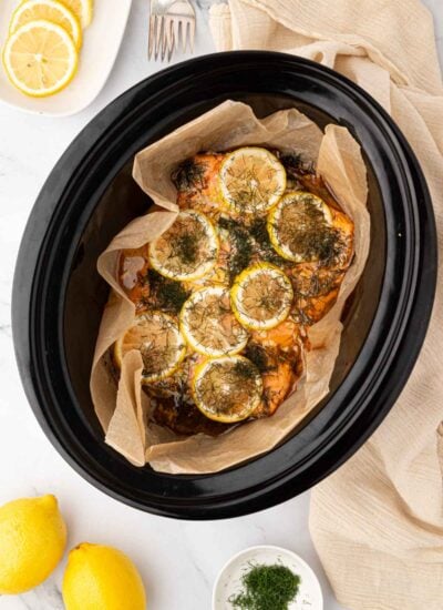 Slow cooker salmon with lemon slices and dill on top in the black bowl of a crockpot. Parchment paper is around the salmon.