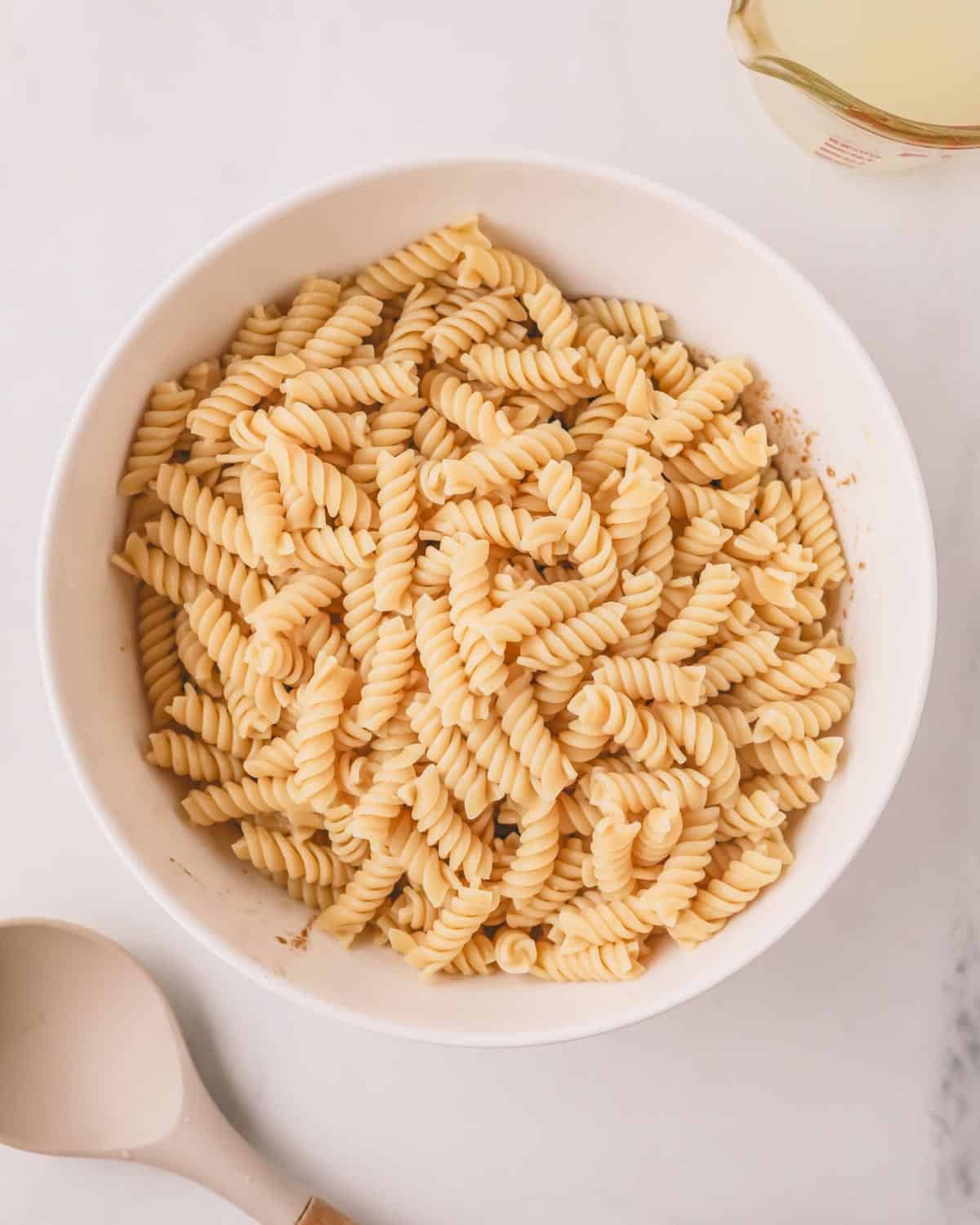 A white bowl with boiled rotini noodles and a gray spatula on the bottom left.